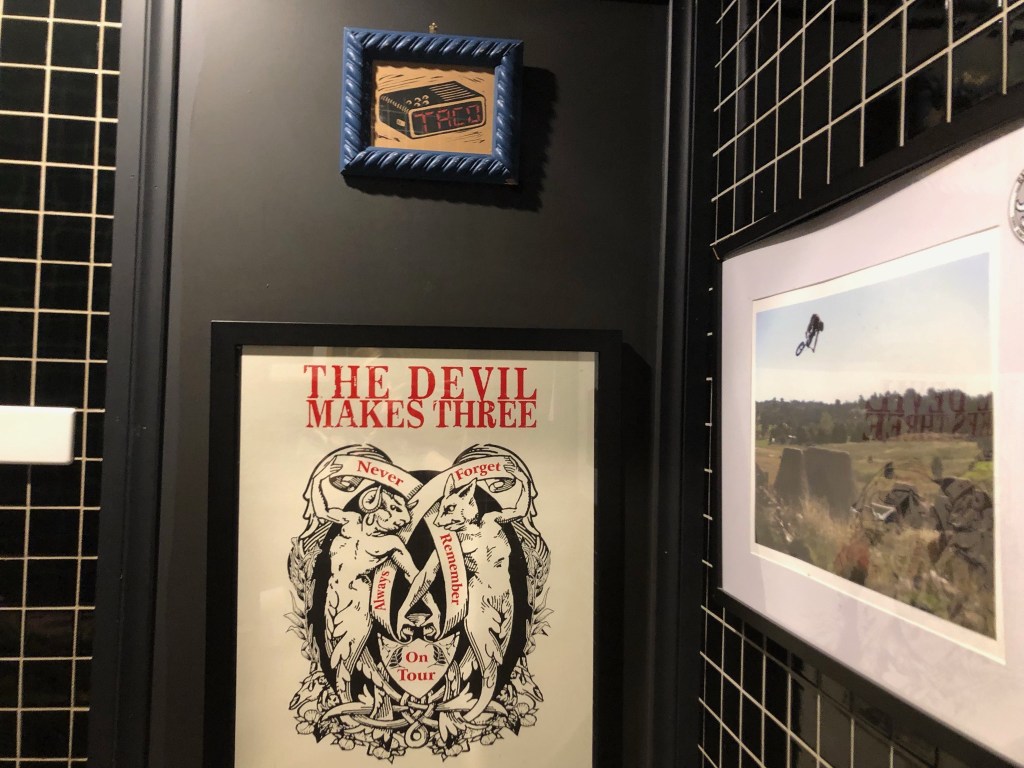 Three images hang on bathroom door with black and white tiles. The first is a small blu framed picture that says the word 'taco.' Below it is a framed images that says 'The Devil Makes Three,' never forget , always remember on tour.' On the side is an image of a BMX riding doing a jump, behind a clear blue sky. 