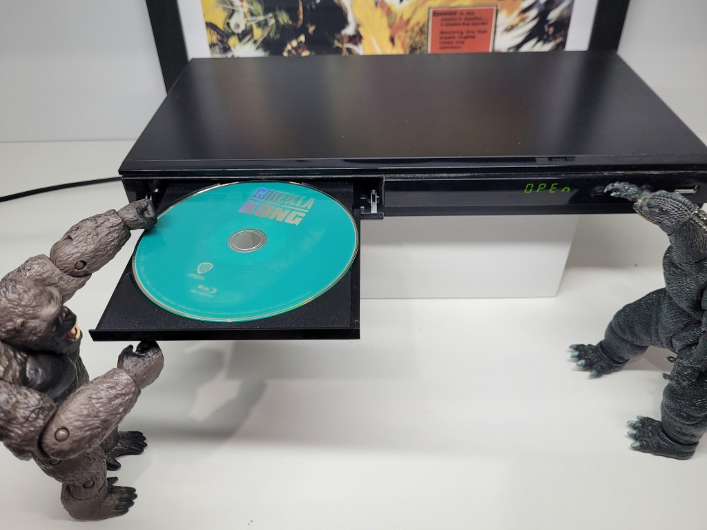 A King Kong figurine feeding the Blu-Ray disc of Godzilla vs Kong into a Blue-Ray player on a white plinth as a Godzilla figurine presses a button to open and close it.