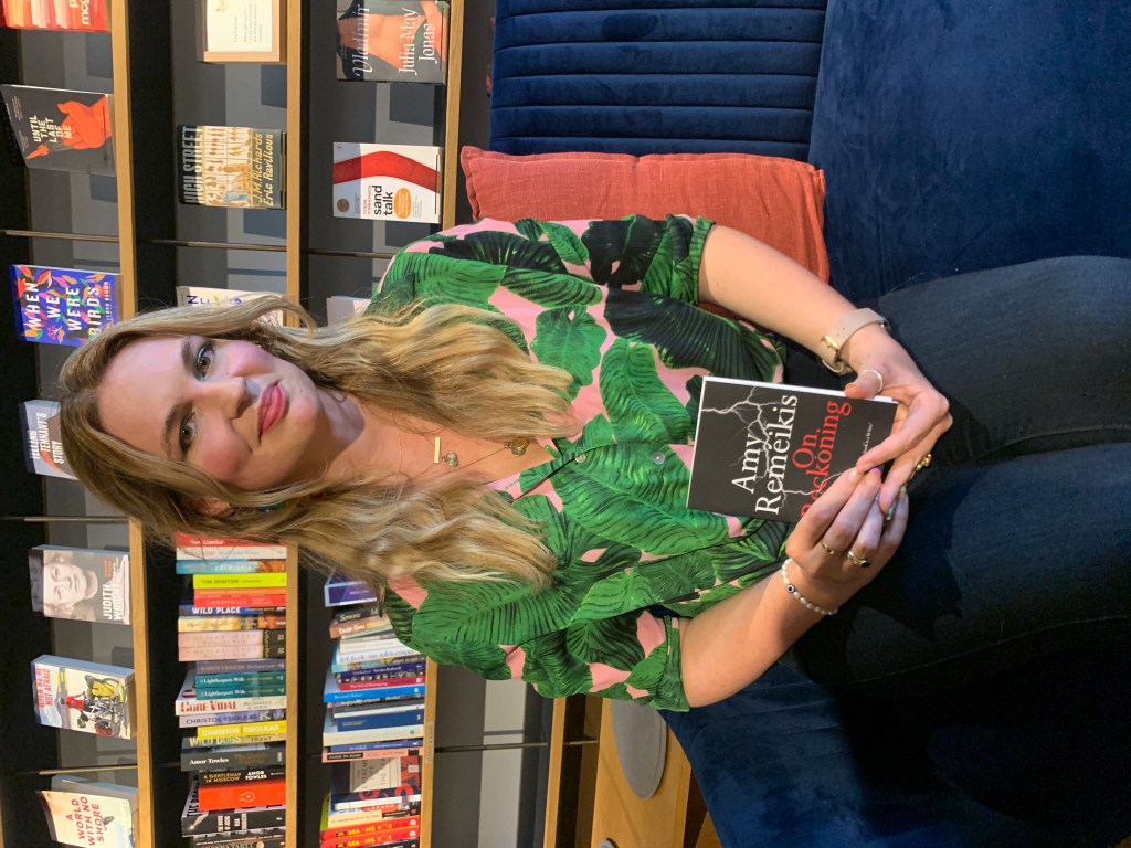 Amy sits on a blue, velvet chair, in front of a wall of books. She holds a copy of On Reckoning, and wears a green and pink shirt with leaves on it. She has blonde hair, and smiles. 