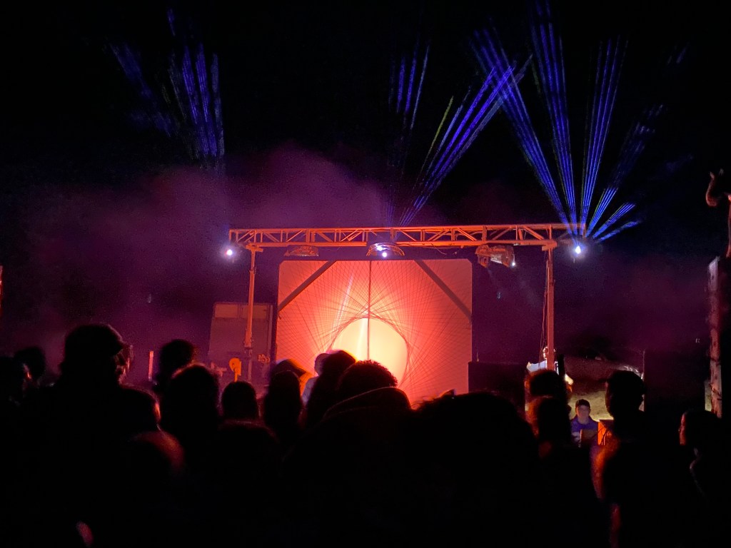 Picture of a stage, backlit red with blue lasers shooting into the night sky, a crowd of people fill the bottom of the image and smoke is seen just above the stage. 