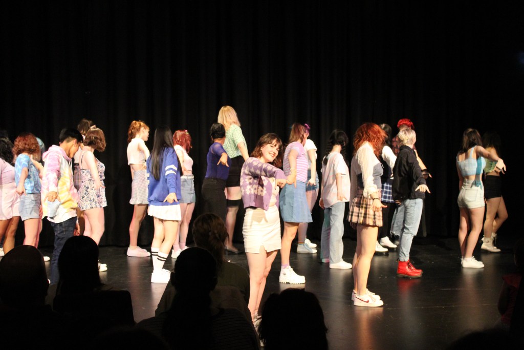 Dancers on the stage, everyone but the girl in the front is facing the back with their hands facing the camera
