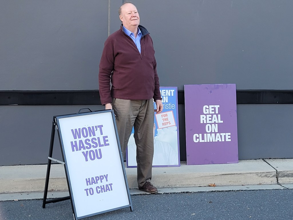 Dr Christie posing next to corflutes that say "Won't Hassle You" and 'Get Real On Climate"