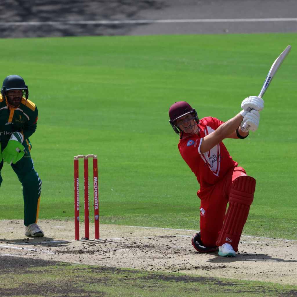 A cricket player in red hits the ball in front of the wicket