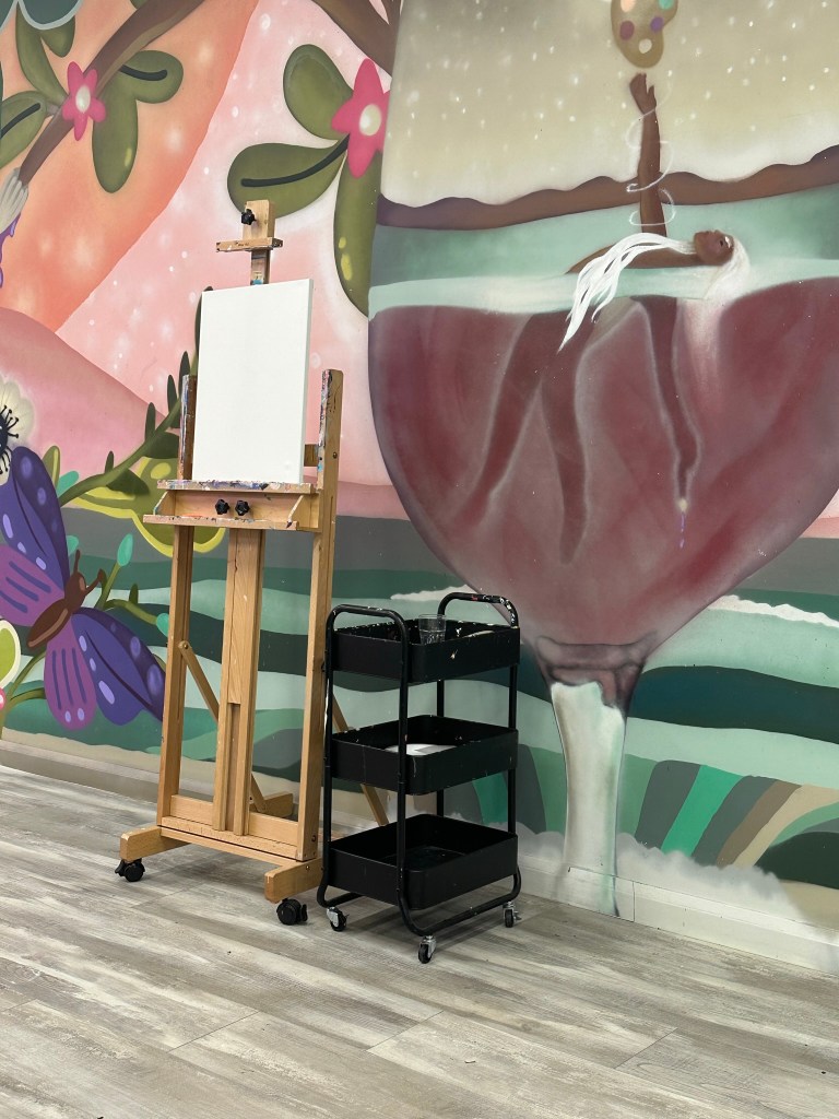 Sitting in front of a large mural painted wall are a plain white canvas on a painting stand and a black cart.