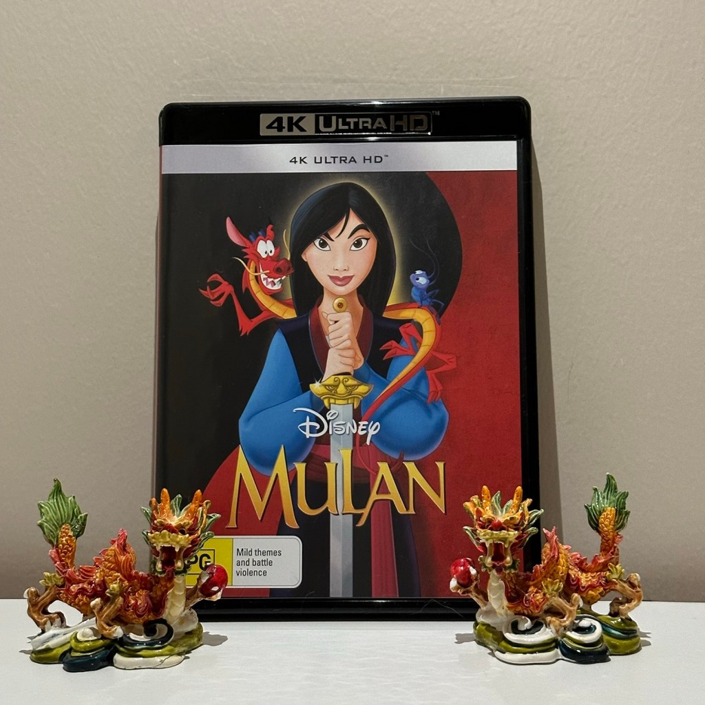 Disney's animated Mulan DVD leaning against a wall with two, small, red dragon statues on either side, framing it.