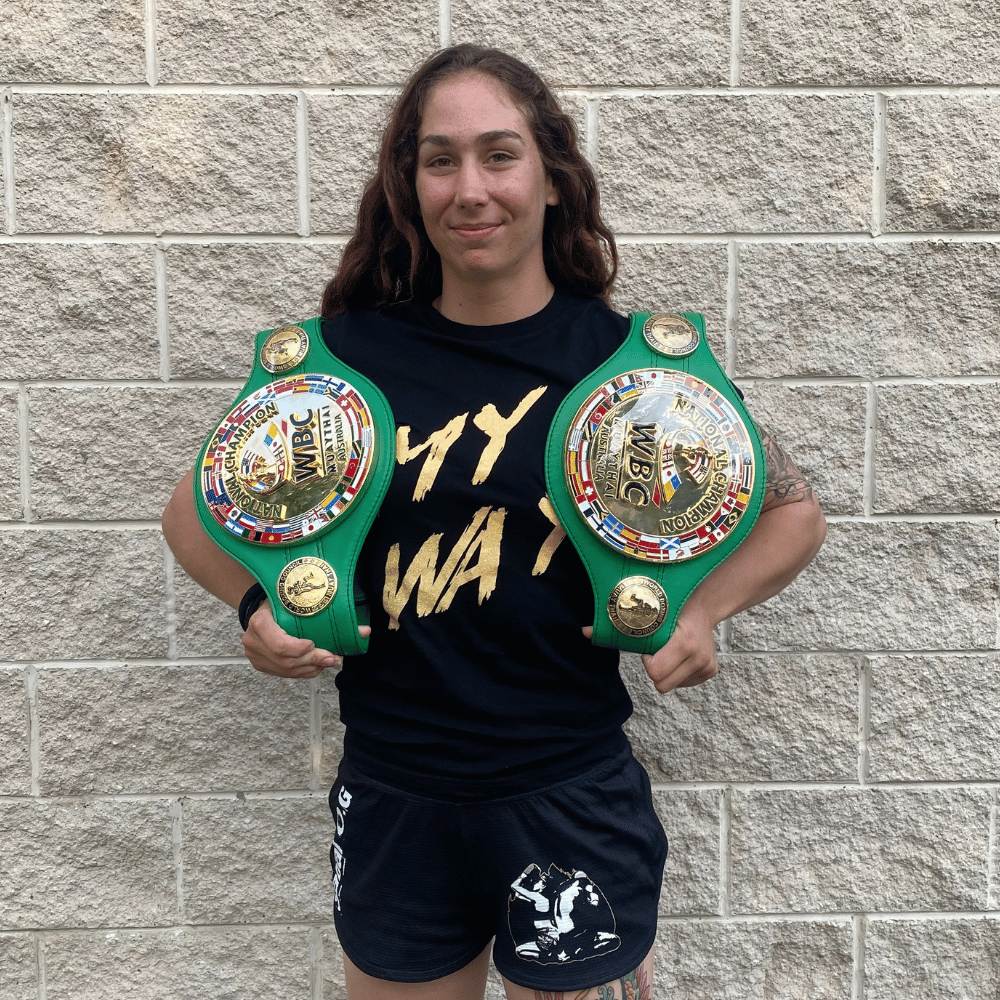 Zoe Putorak holding her two WBC National champion muay thai belts, wearing her signature shirt with her fight name 'My Way' and her team Sabai Muay Thai & Boxing's logo on her shorts. 