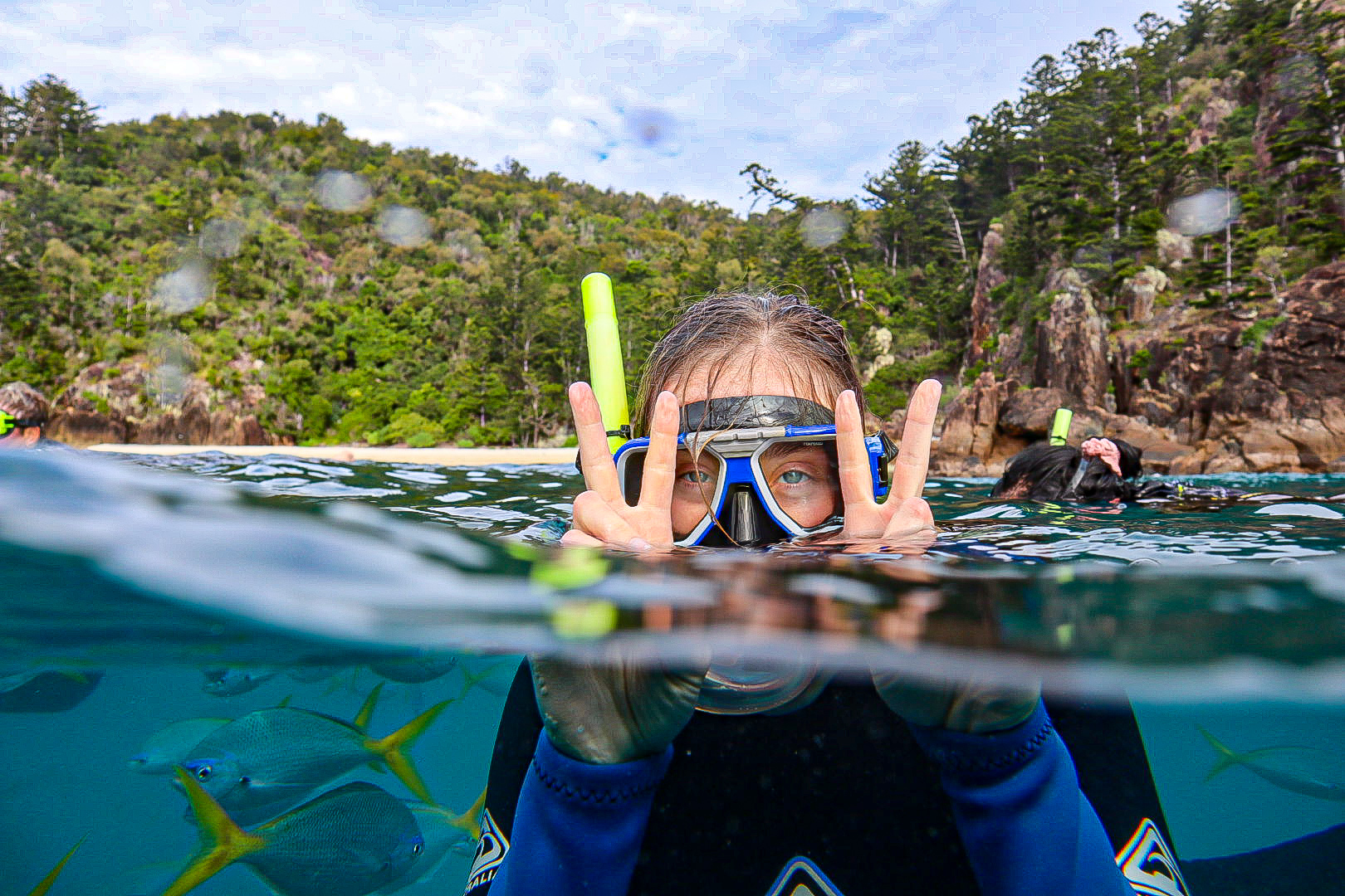 Person in ocean swimming, with snorkelling gear. Holding up the peace sign. Fish below the person. Land, trees and rocks above the person