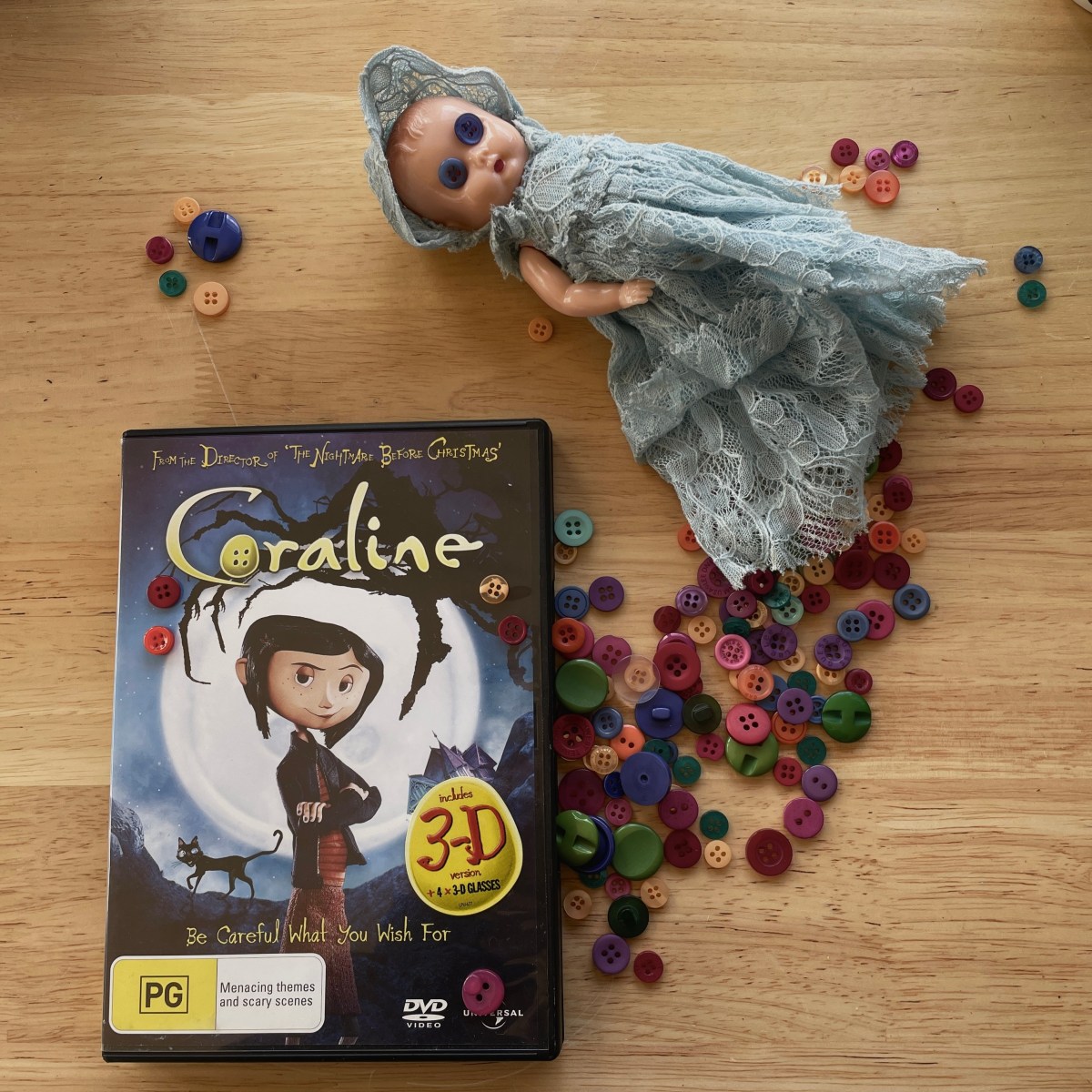 A Coraline DVD lies on a wood bench, on the cover is an animated girl with blue hair and her arms folded. Colourful buttons are spilling out of the DVD case and underneath a small doll with a blue dress and blue buttons for eyes.