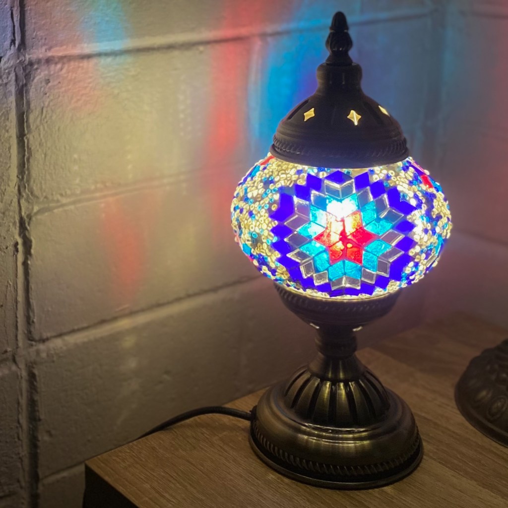 Lamp turned on, sitting on wooden surface, reflecting colour onto white wall. The lamp has a flower pattern with the colours (from inside to out), red, light blue, mirrored and dark blue.  