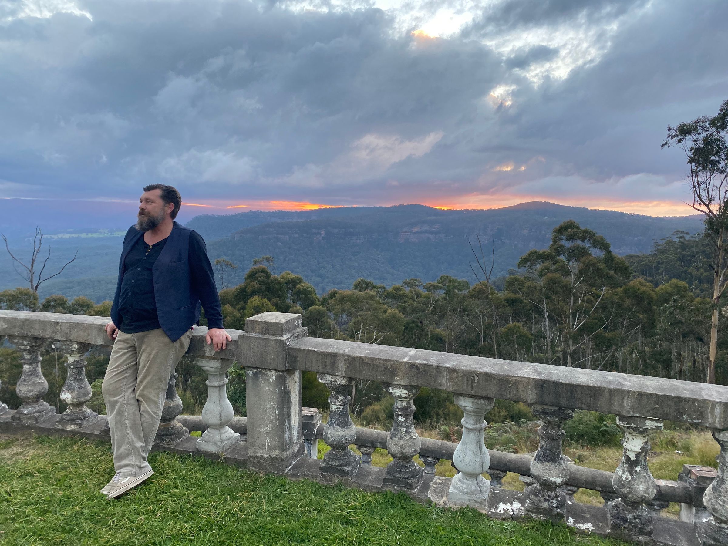 A man is wearing cream pants and a navy shirt with a blue waistcoat and is standing in front of a sunset in the blue mountains where there are a lot of trees.