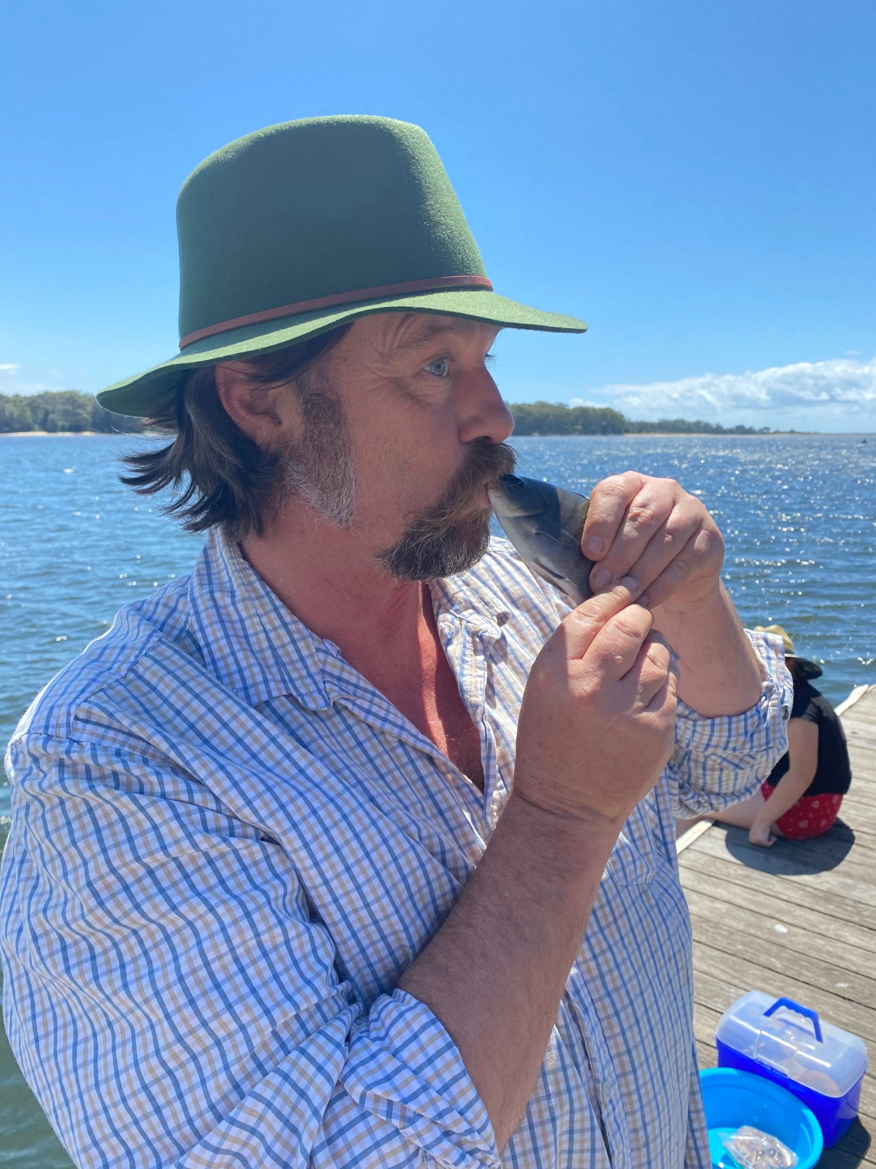 A man wearing a green hat and a white button-up shirt is holding a fish to his lips in front of an ocean. 