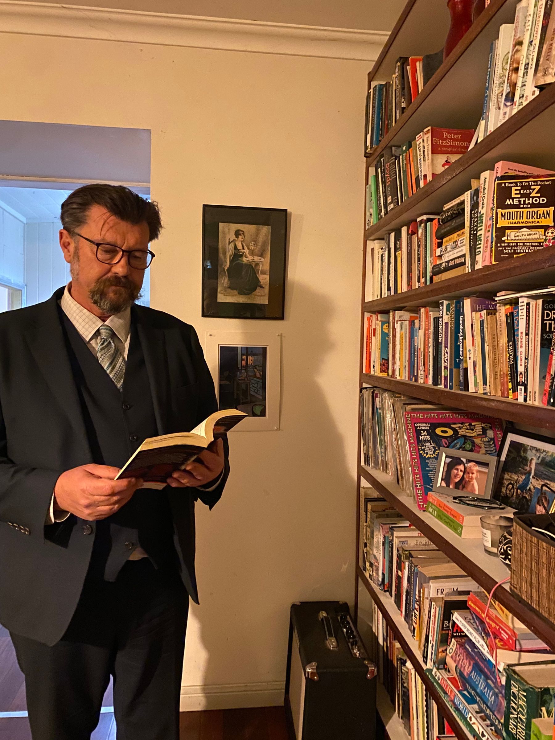 A man is standing in front of a book case and is holding a novel wearing a suit. he is wearing glasses.