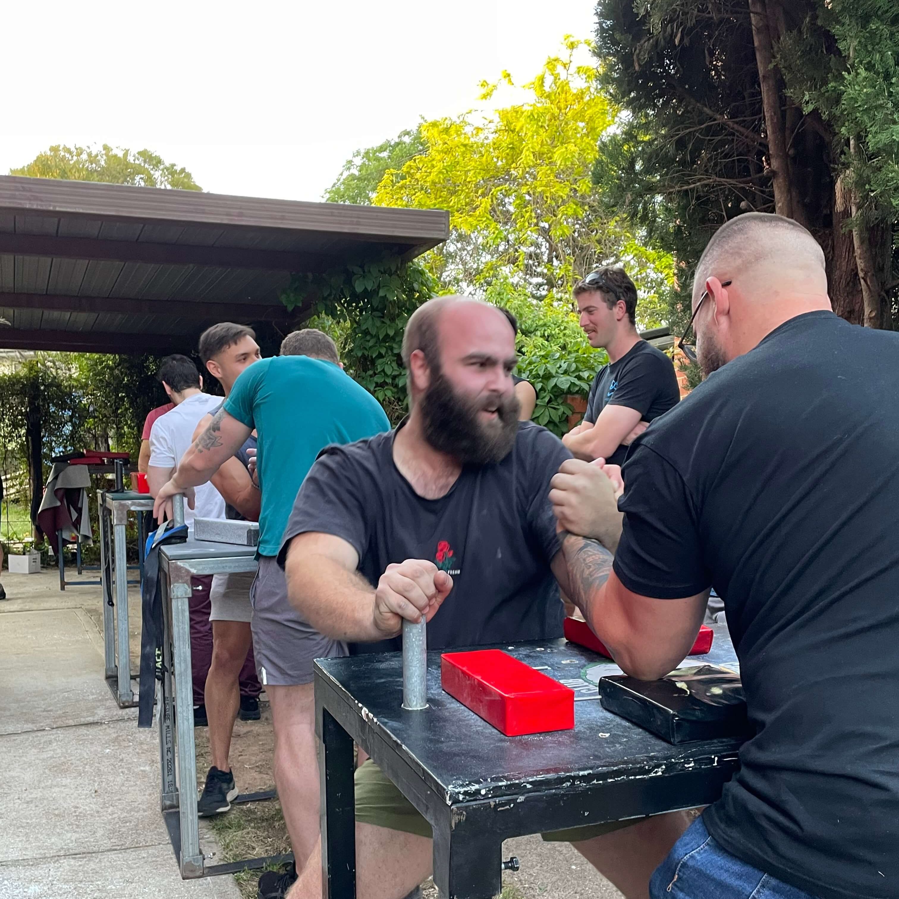 A strong man with a full beard is locked in an arm wrestling match. He is holding onto the table, and is clearly putting the effort in