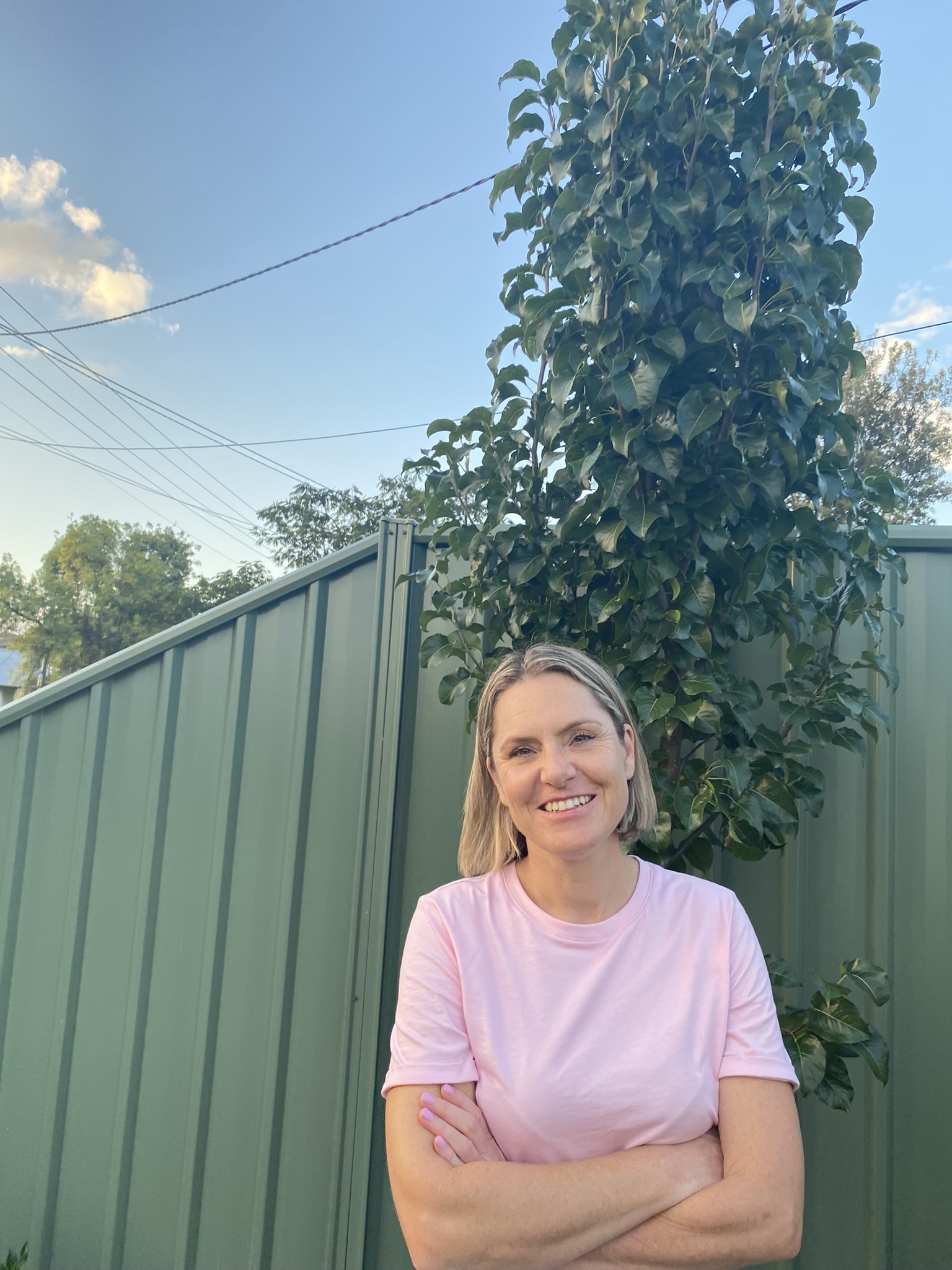 An image of a Mel. She has short blond hair and a pink-shirt, she is smiling towards the camera. She stands in front of a green fence and tree within a suburban Australia neighborhood.