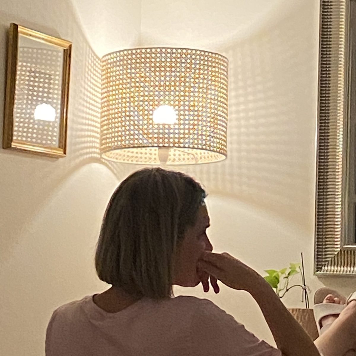 A short-haired woman sits at a table with a contemplative pose. A lamp just above her illuminates a halo-like light.