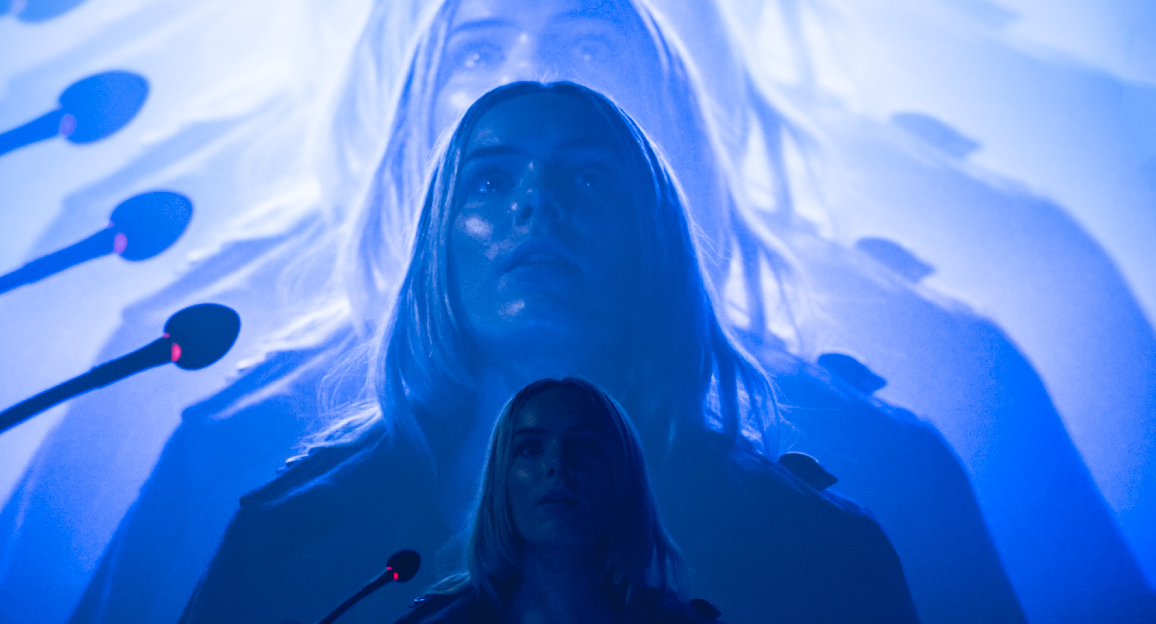 Image of a woman standing in front of a microphone stand with repeating projection highlighted in blue being projected behind her. 