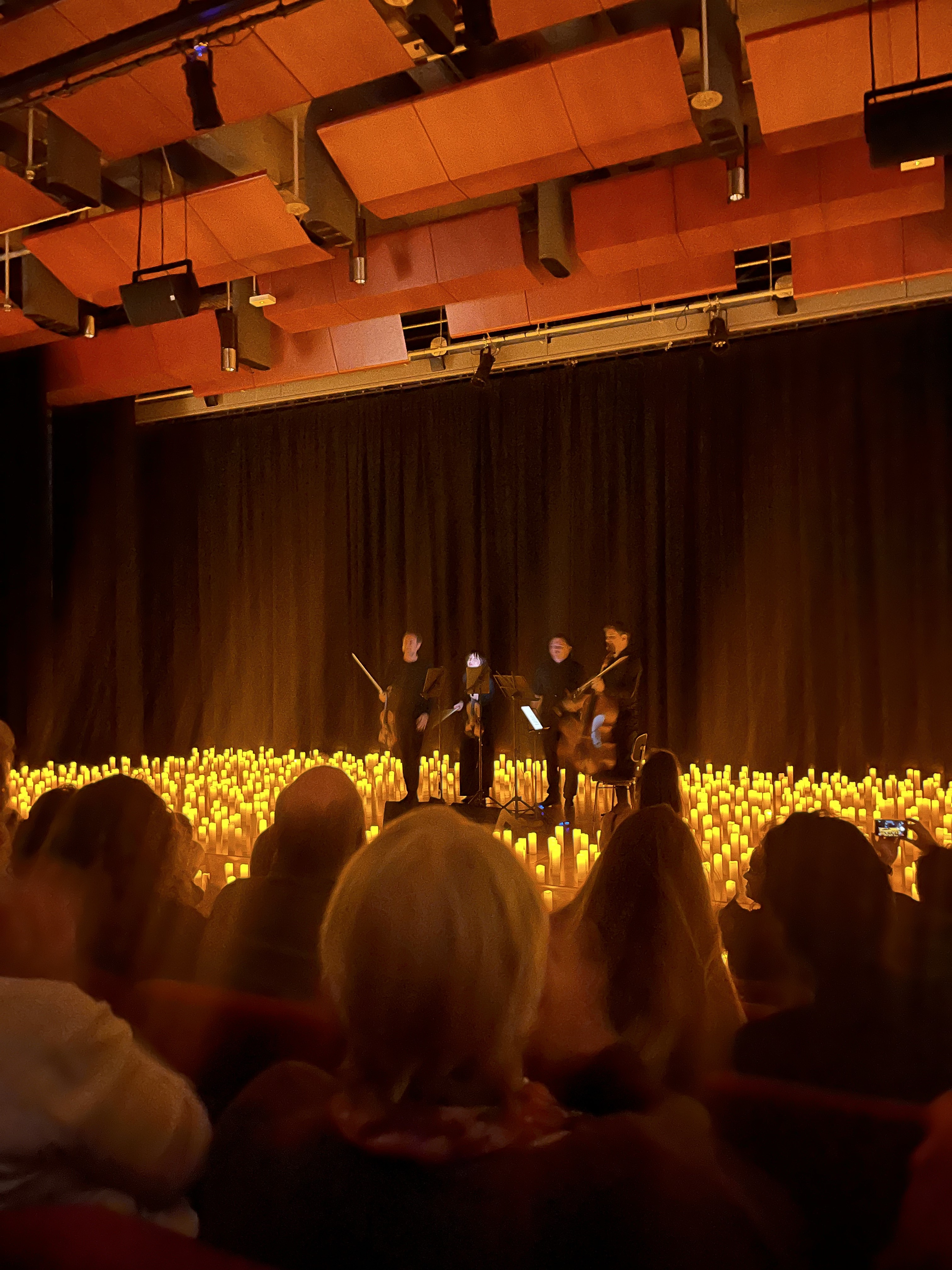 Four people playing a string instruments bow on a stage covered in candles as audience watches.