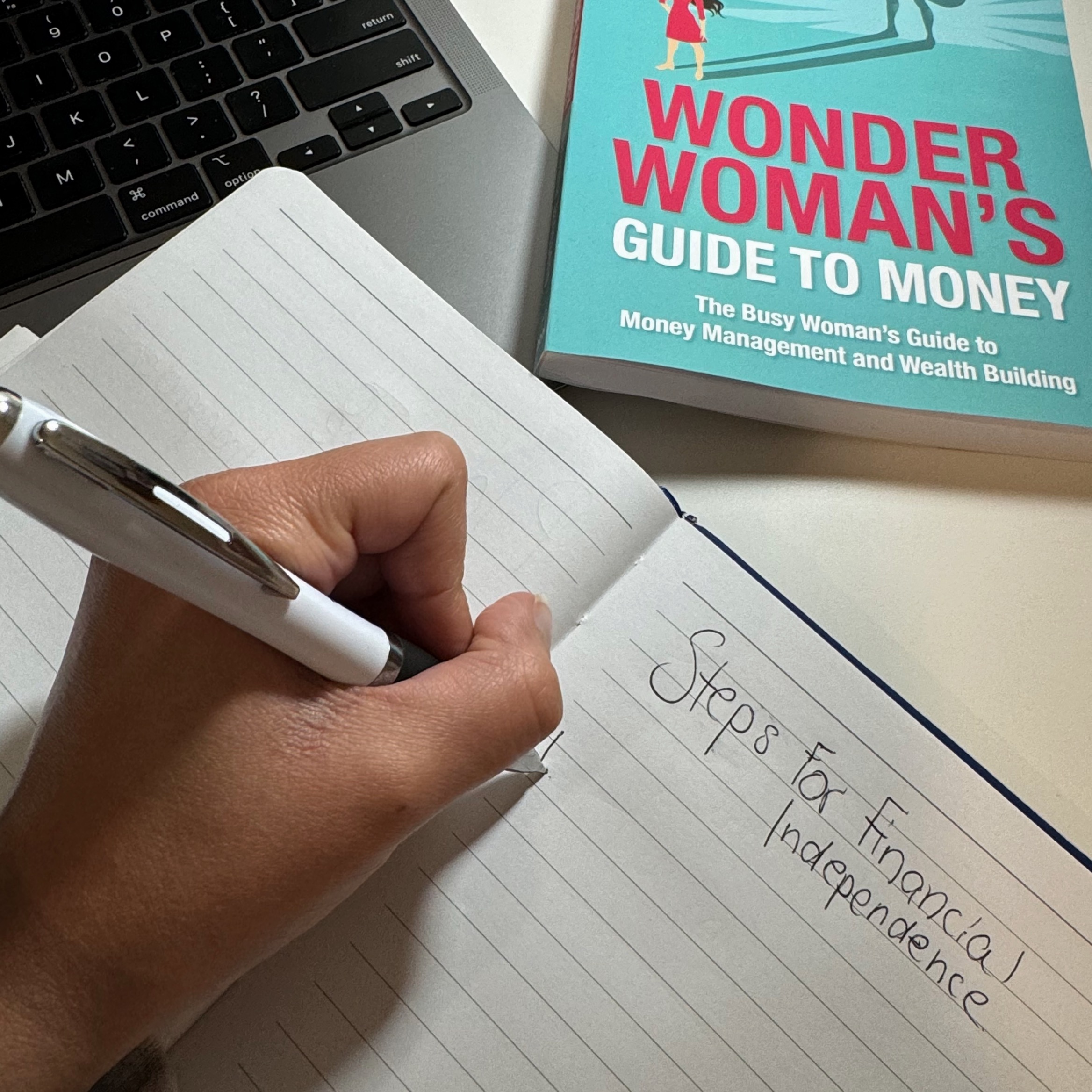 person writing in a notebook with book and computer in the background 
