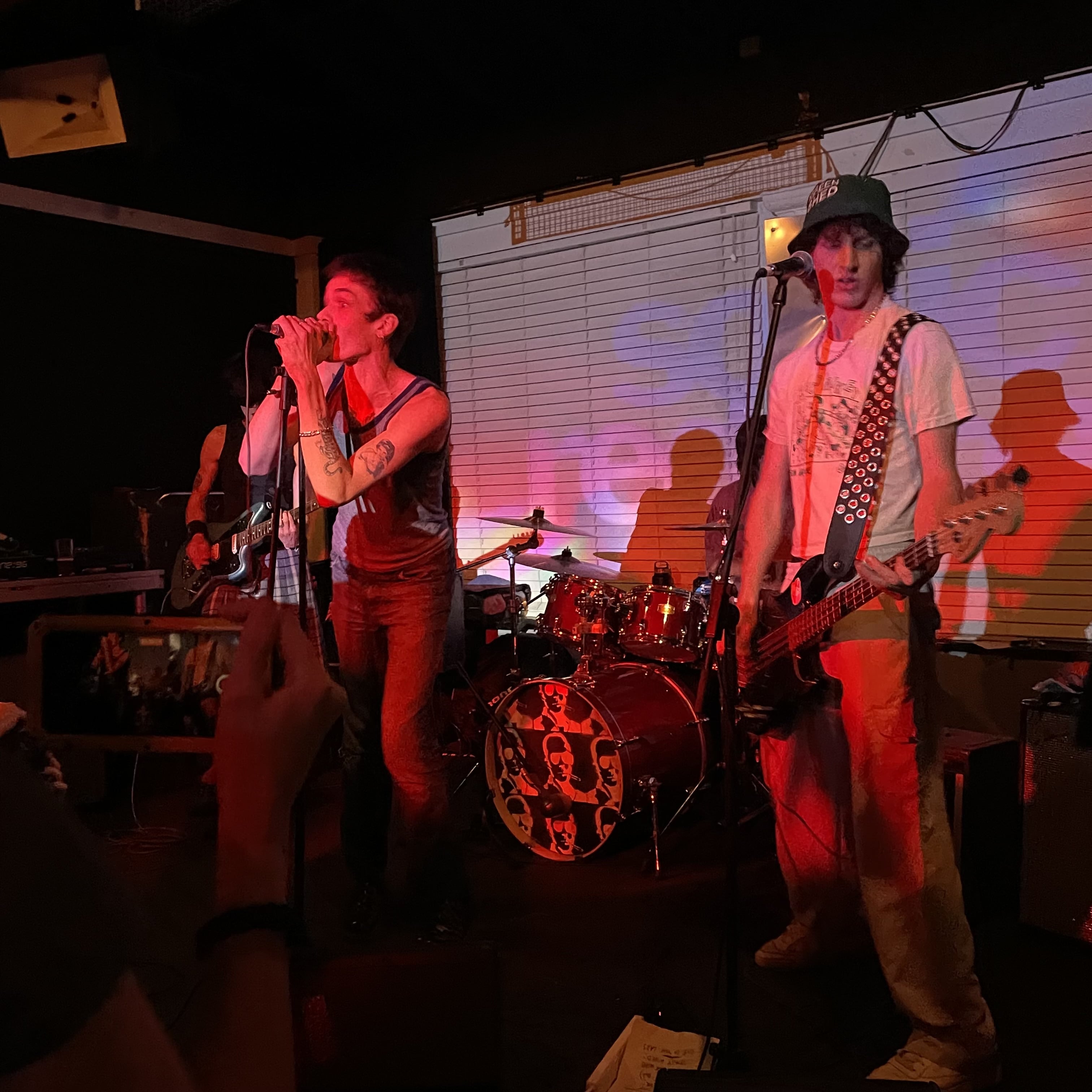 Punk band Sonic Reducer on stage. The singer grips the microphone, standing to the left. The bassist plucks along, standing to the right. A red light is shrouded on them
