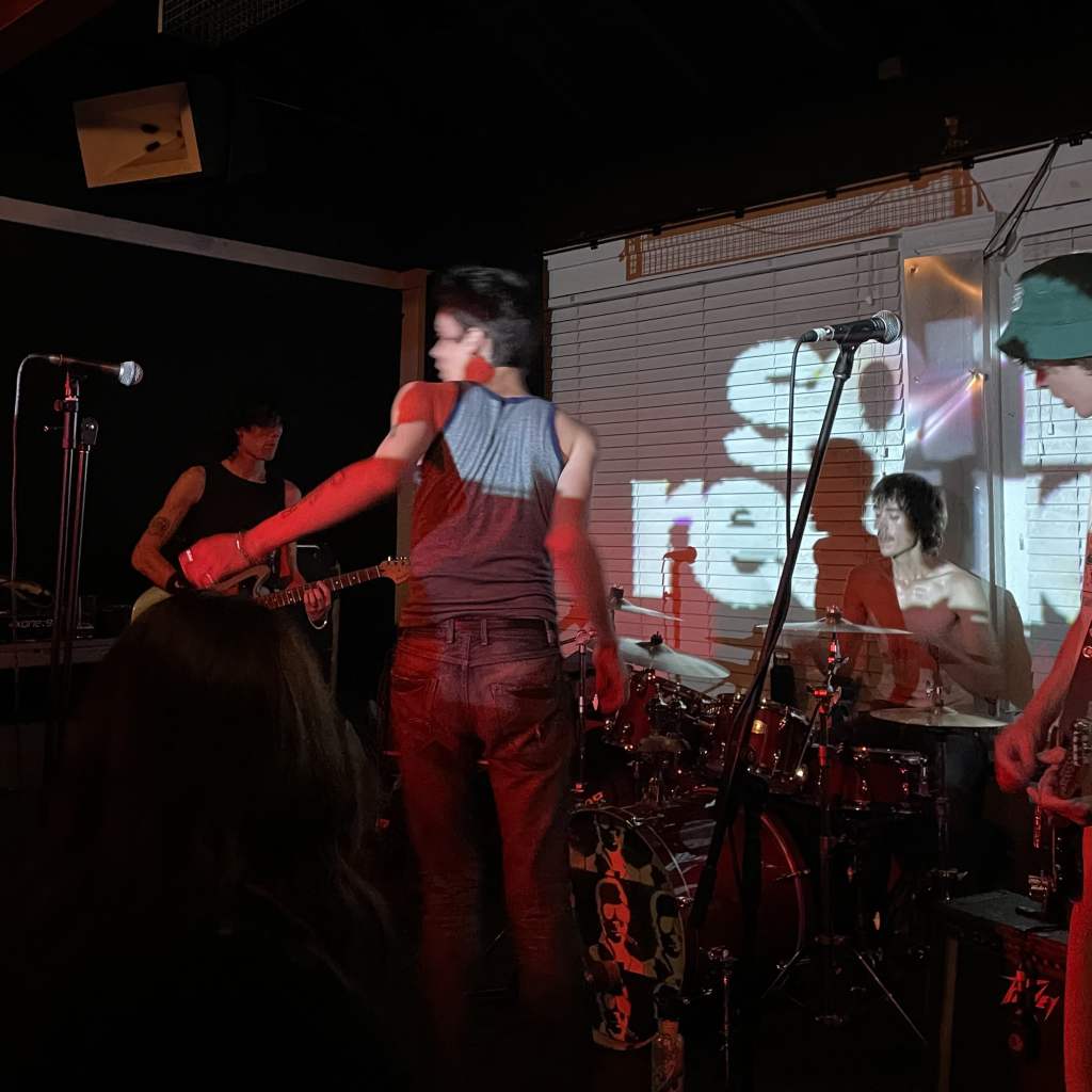 Punk band on stage. The singer is moving around with his back to the camera. He's about to spin around. The drummer and guitarist are sweaty and steady.