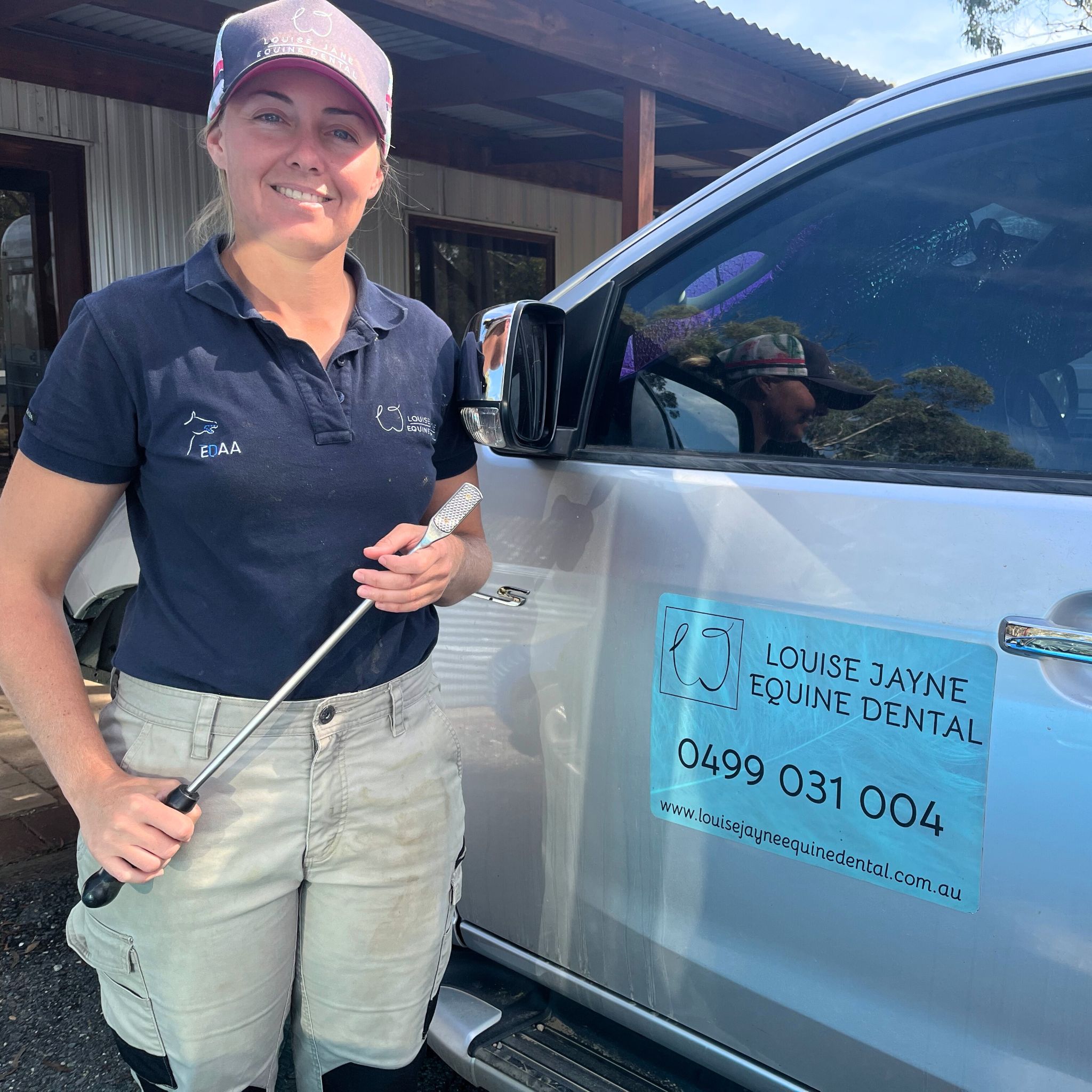 A woman wearing a baseball cap, polo shirt and white pants holds a titanium file used for filing horse's teeth. She is standing next to a car with a sign on the side that reads 'Louise Jayne Equine Dental'.