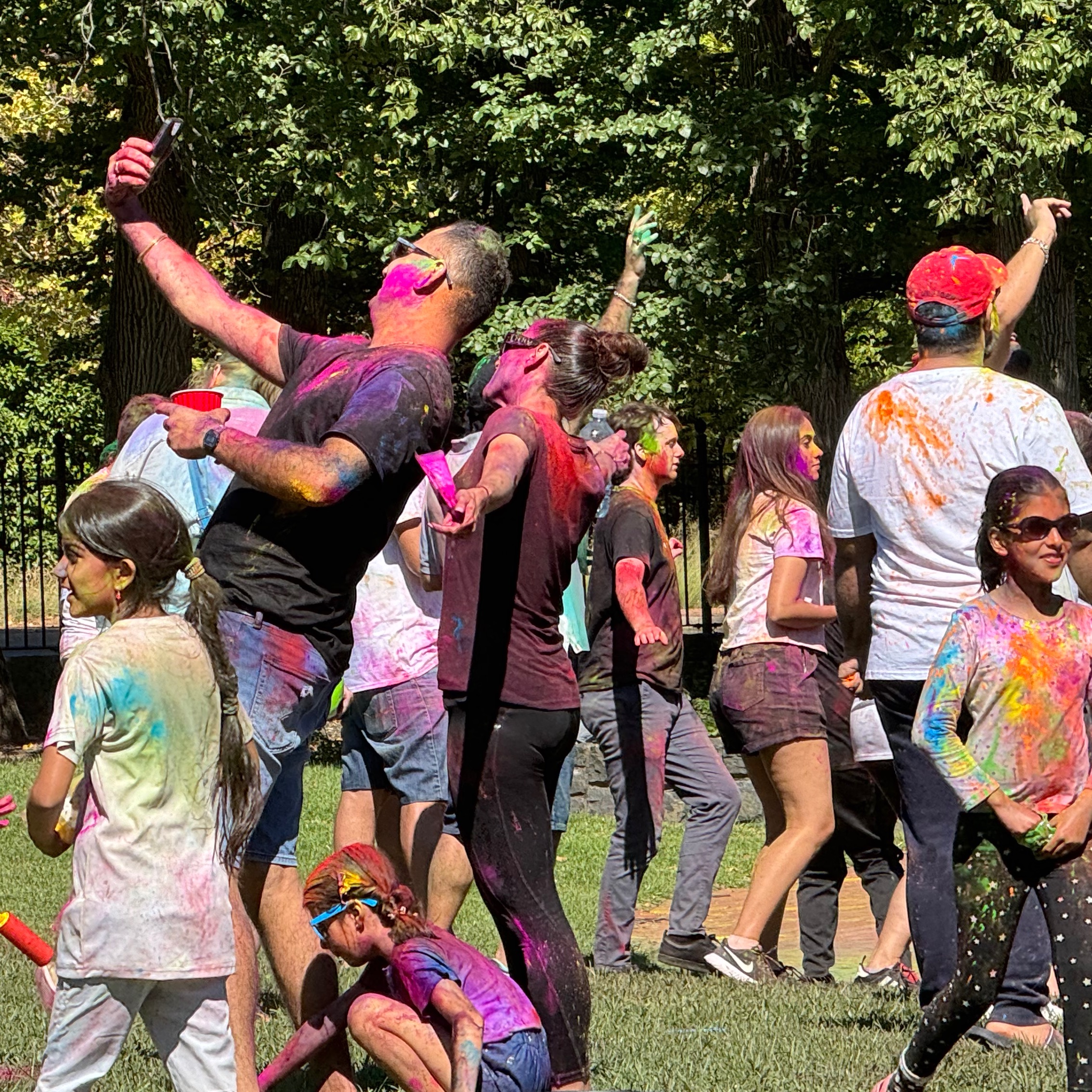 woman and man taking a selfie. People dancing in the background. Girl wearing sunglasses with hand in packet of green colour. Kids bending down. 