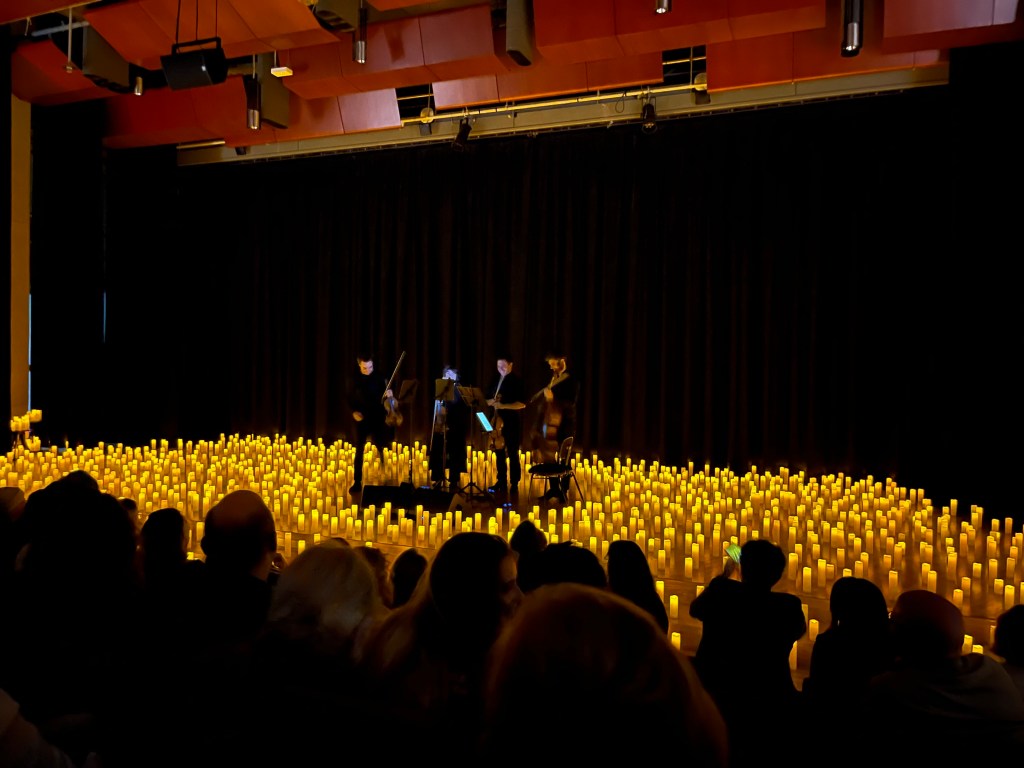 Four people playing a string instruments perform on a stage covered in candles as audience watches.