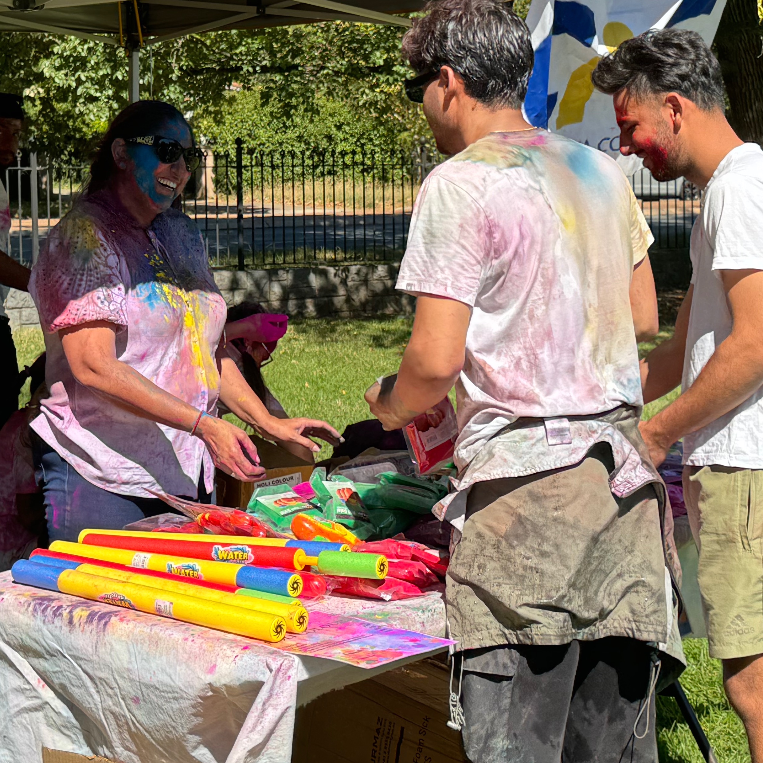 woman selling water guns and packets of colour at a stall to two men. 