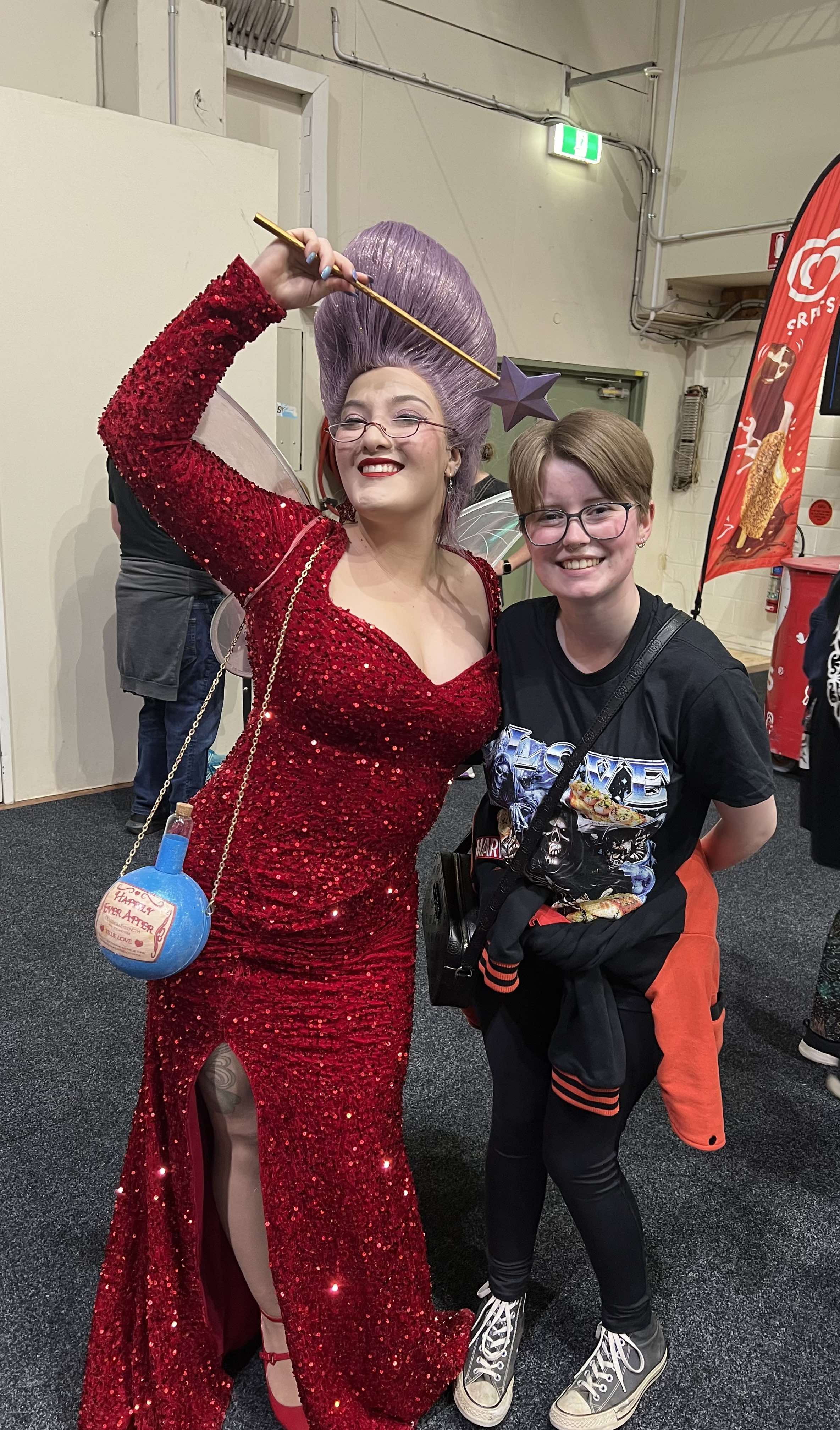 Amelia Torresan is dressed as the Fairy Godmother from the Shrek films. She has a high light purple, sparkly wig on, wearing glasses ad bright red lipstick. She has a form fitting sparkly red dress on, with a sweetheart neckline. The dress also has a high slit on right side, showing her leg, red high heels and half of her thigh - her thigh also showing a partial tattoo. On the right side of her body she is wearing a potion bottle bag, with blue potion inside - the text on the front of the bag says, Happily Ever After. She is also posing with her right arm up, holding a gold wand with a purple star on the end - the purple is the same shade as her wig. Azzie has a black tshirt on, with a design on the front, they has a black cross body bag, and black leggings on. They also have a black and red jacket tied around their waist. Their shoes are grey converses.