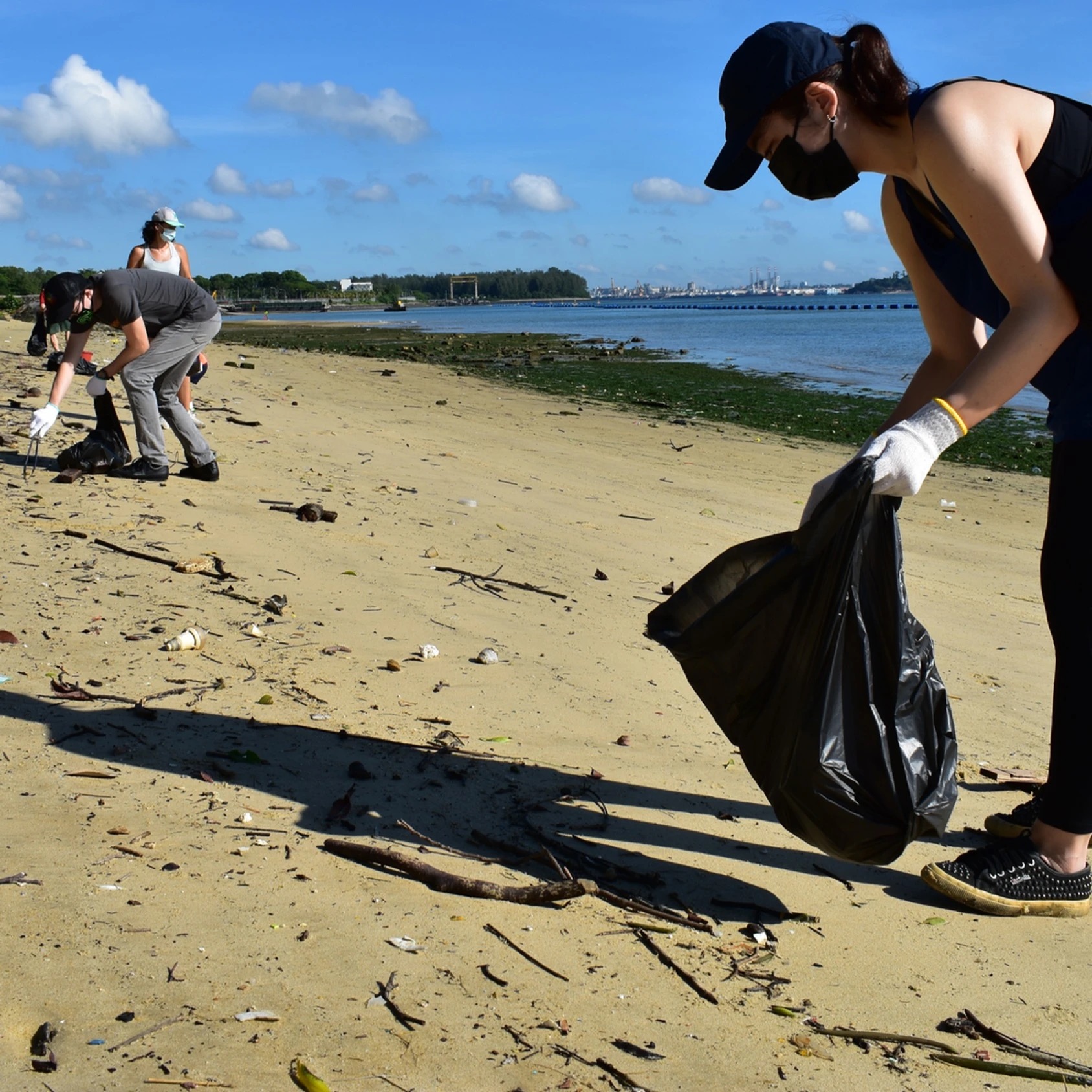 woman wearing hat and a mask holding trash bag and cleaning the beach, man in background holding tongs to pick up dirt off the sand and woman in the background standing behind the man on the beach 
