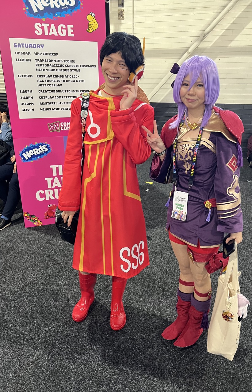 Jeff cosplaying as Luffy from One Piece. He is wearing a short black wig, a red and yellow ear piece, a red dress with gold stripes running down the front. The left side of the dress has SSG on the bottom, he is wearing red boots as well as a black shoulder bag with various different pins.
Nina is cosplaying as Bernadetta Von Varley from Fire Emblem. She has a light purple wig on, with a black bow in the right side. Her dress is purple with a gold design on front (two gold lines on the front with swirling design under the lines. On the top is a gold cape, with a red shoulder pads on top. On the left side of the dress, there is a red and gold diamond design and a gold design with a thick gold line under it. She also has a red shorts under the dress, with red shoes and a purple accent on top). Nina is carrying a light colour tote bag, with two bag charms. Nina is also wearing a media pass around her neck, and posing with a piece sign.