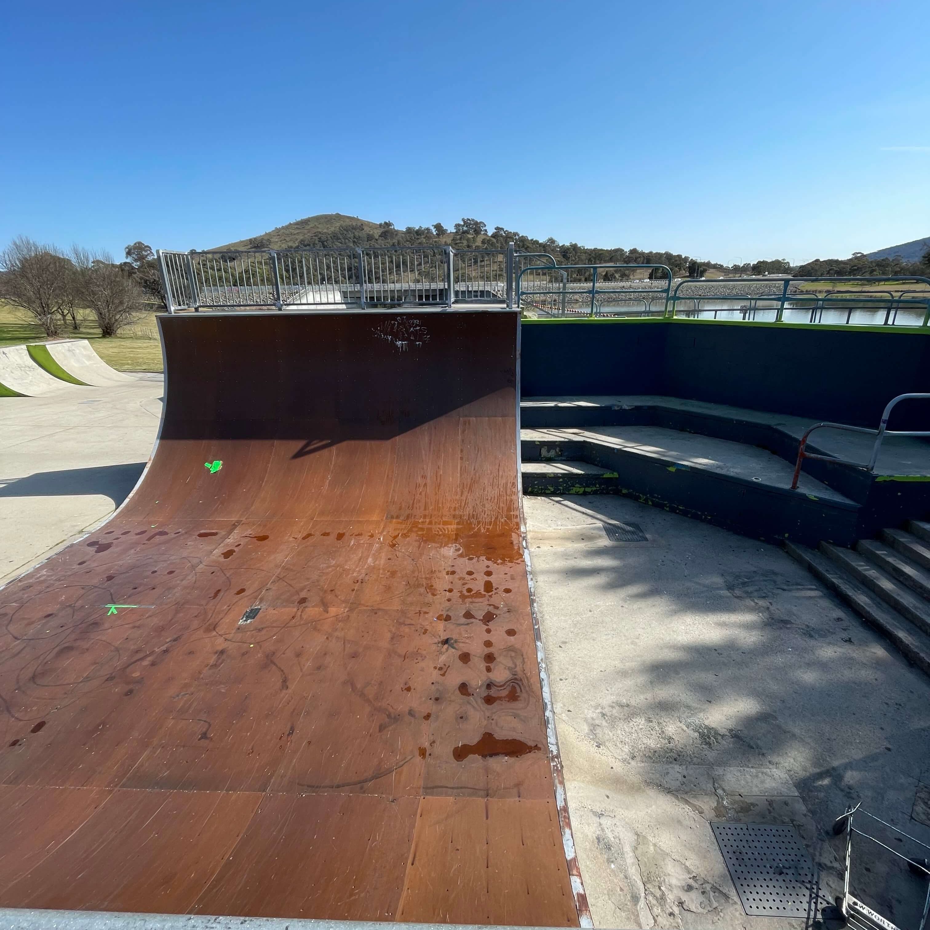 A smaller, dirty, dingier looking vert ramp at the Tuggeranong skatepark. It looks worn out, with little puddles on the ramp. 