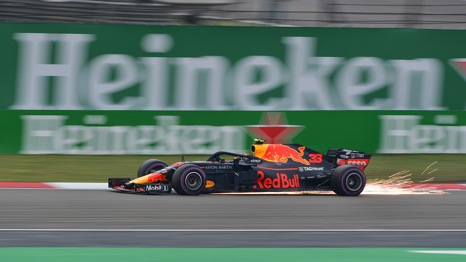 A black F1 car with a yellow and red bull on the top and front, is driving on a track. There are sparks flying out the back of the car. The number 33 can be seen on the side as well as Red Bull in bold red letters. This is Max Verstappen's 2018 car. The driver himself, cannot be seen in the image. Behind the car, out of focus, is the Heineken logo.