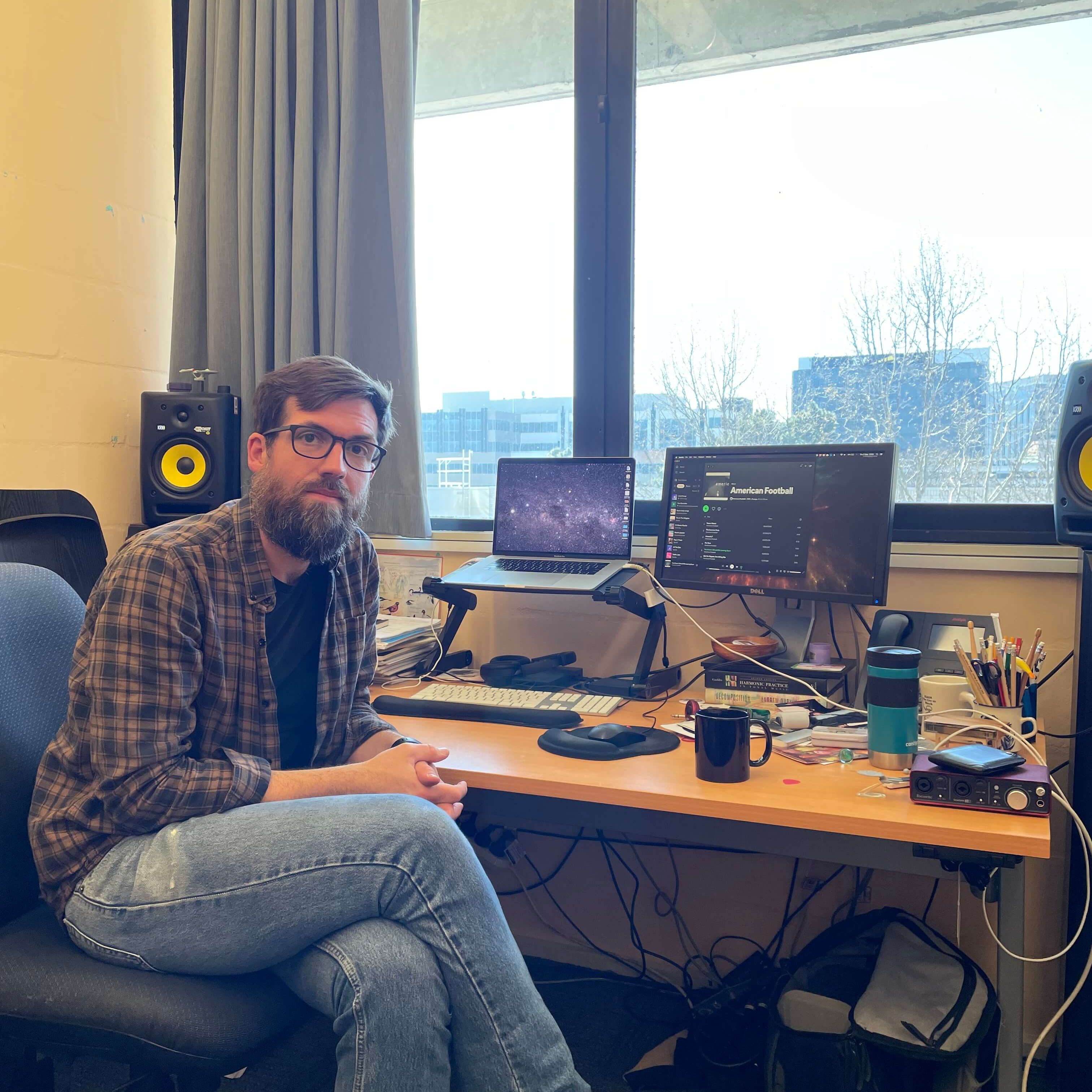 A man with a beard and glasses sits at his desk, looking towards the camera. On his computer screen he has the album discussed in this article. His desk is cluttered with various knick-knacks.