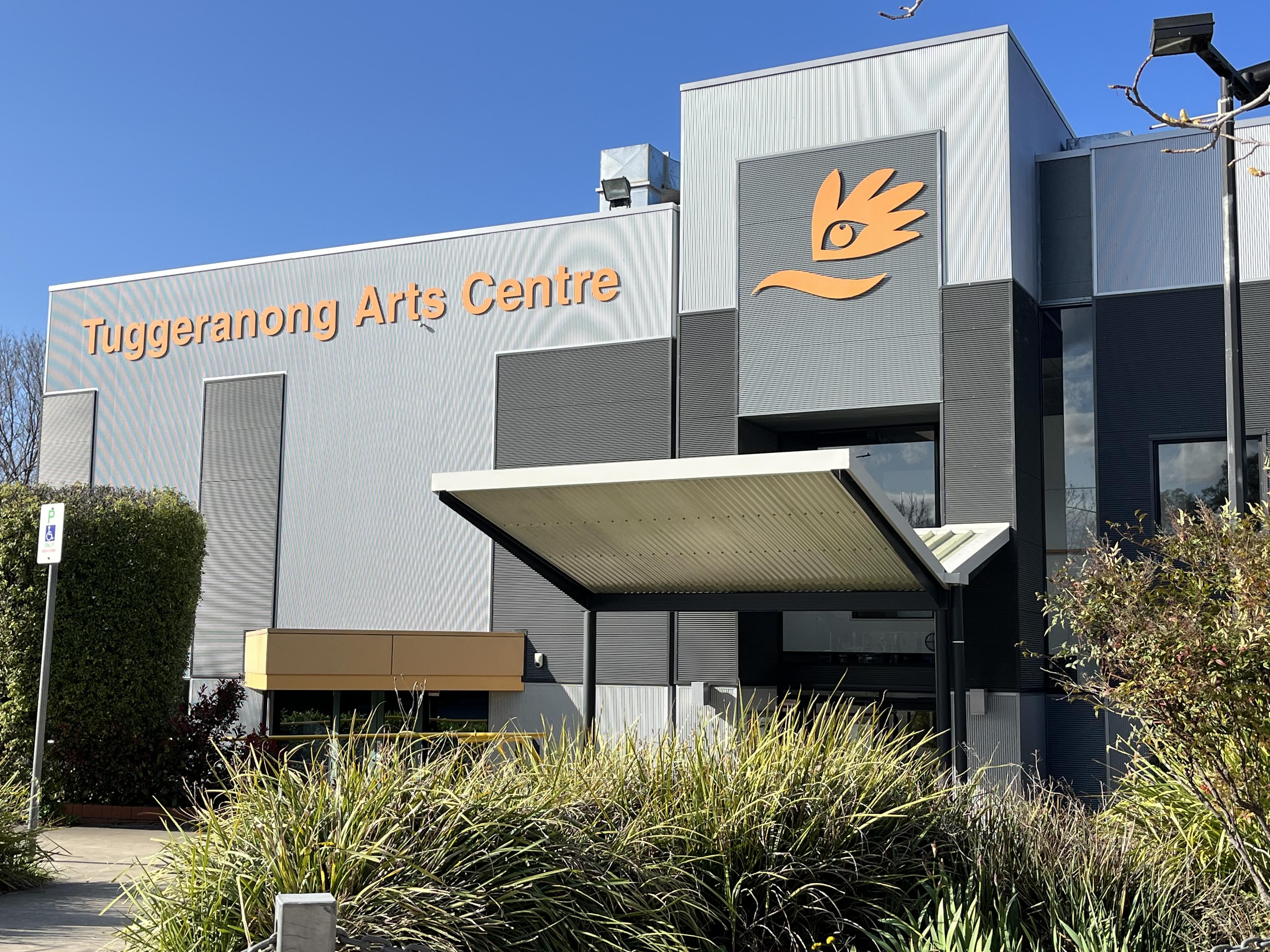 Yellow Tuggeranong Arts Centre sign on big grey building in front of blue skies.