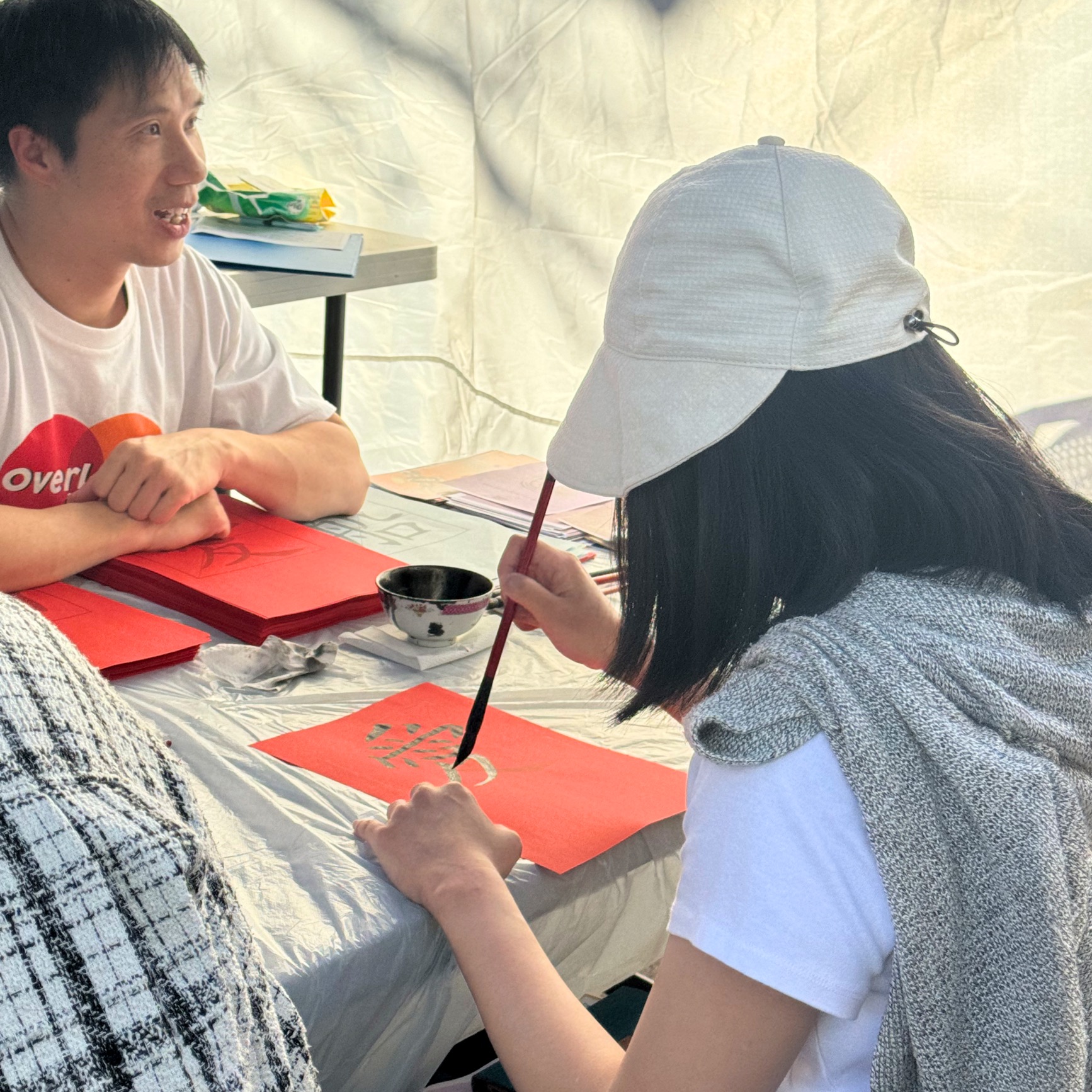 woman wearing jacket and cap holding paintbrush drawing calligraphy with a man with folded arms sitting across from her and talking on a table with a white cloth  and red papers and another person sitting next to the woman