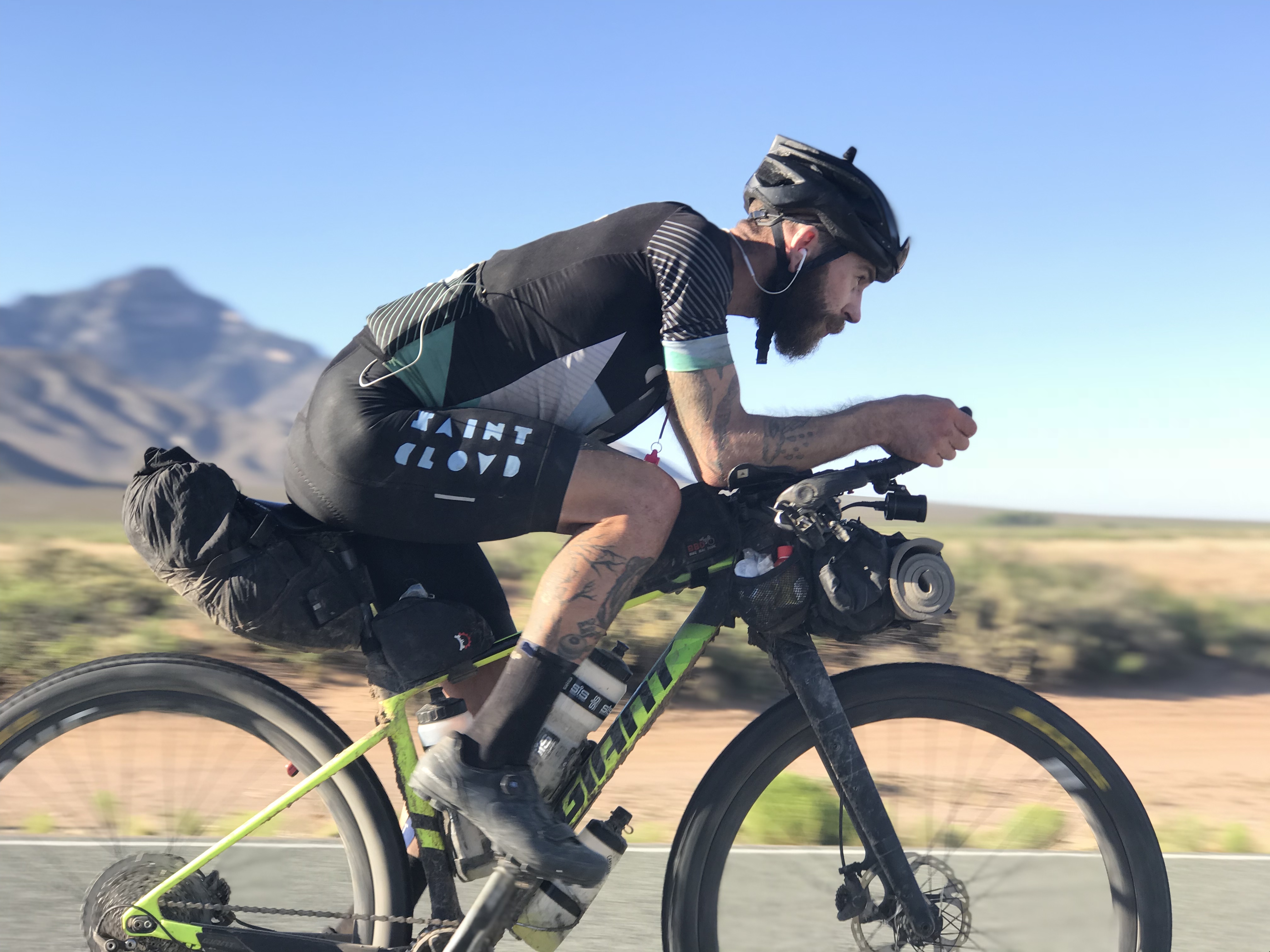 A photo of Lewis Ciddor, a bearded man dressed in exercise gear, cycling on a green mountain-bike along a paved road. The background contains a dusty plain with sparse bushes and a mountain in the distance.