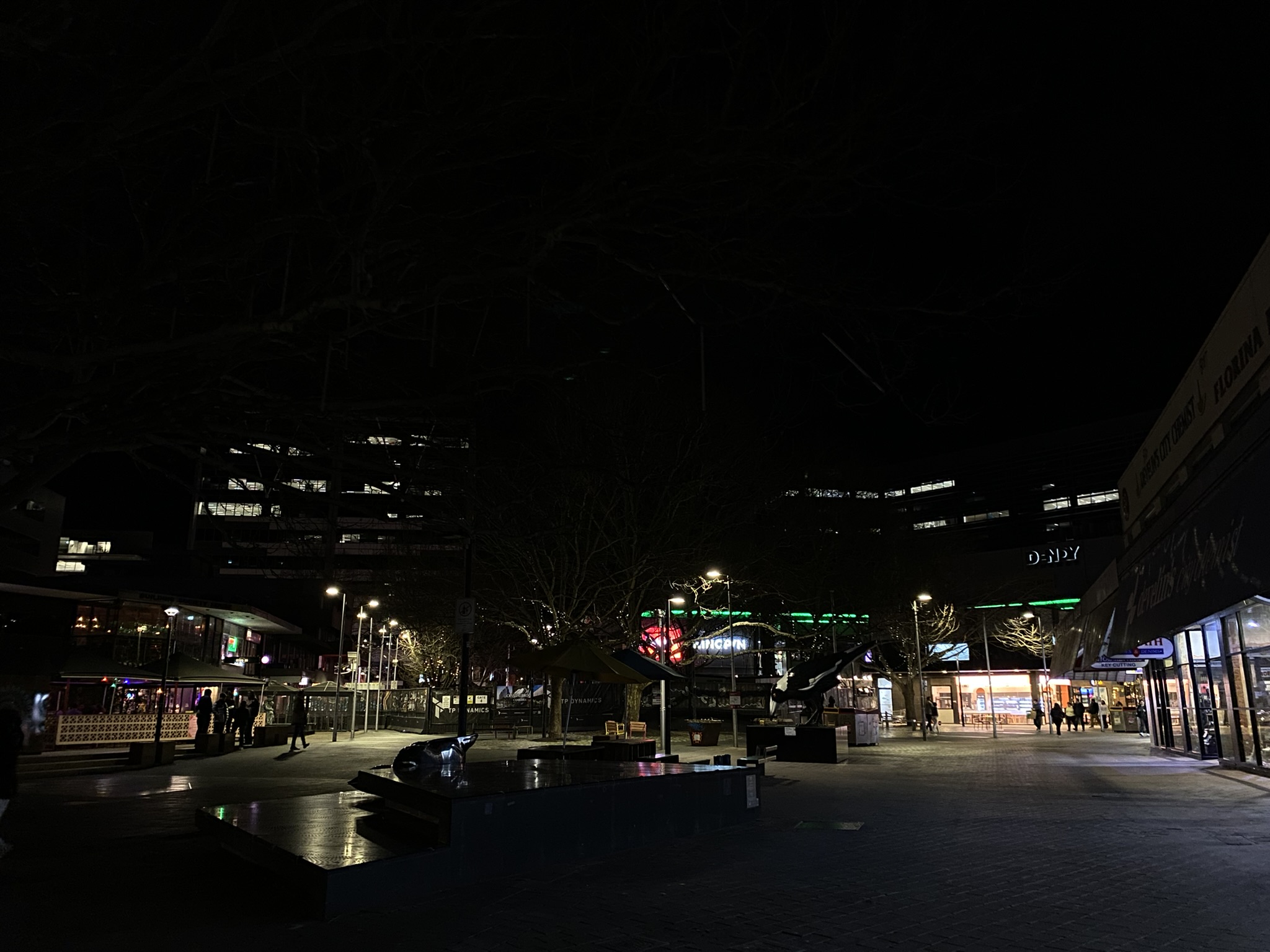 A landscape photo of Canberra's CBD at night, a common area is illuminated by various streetlights in the center with light also illuminating from nearby storefronts and buildings. The sky is completely black with no visible stars.