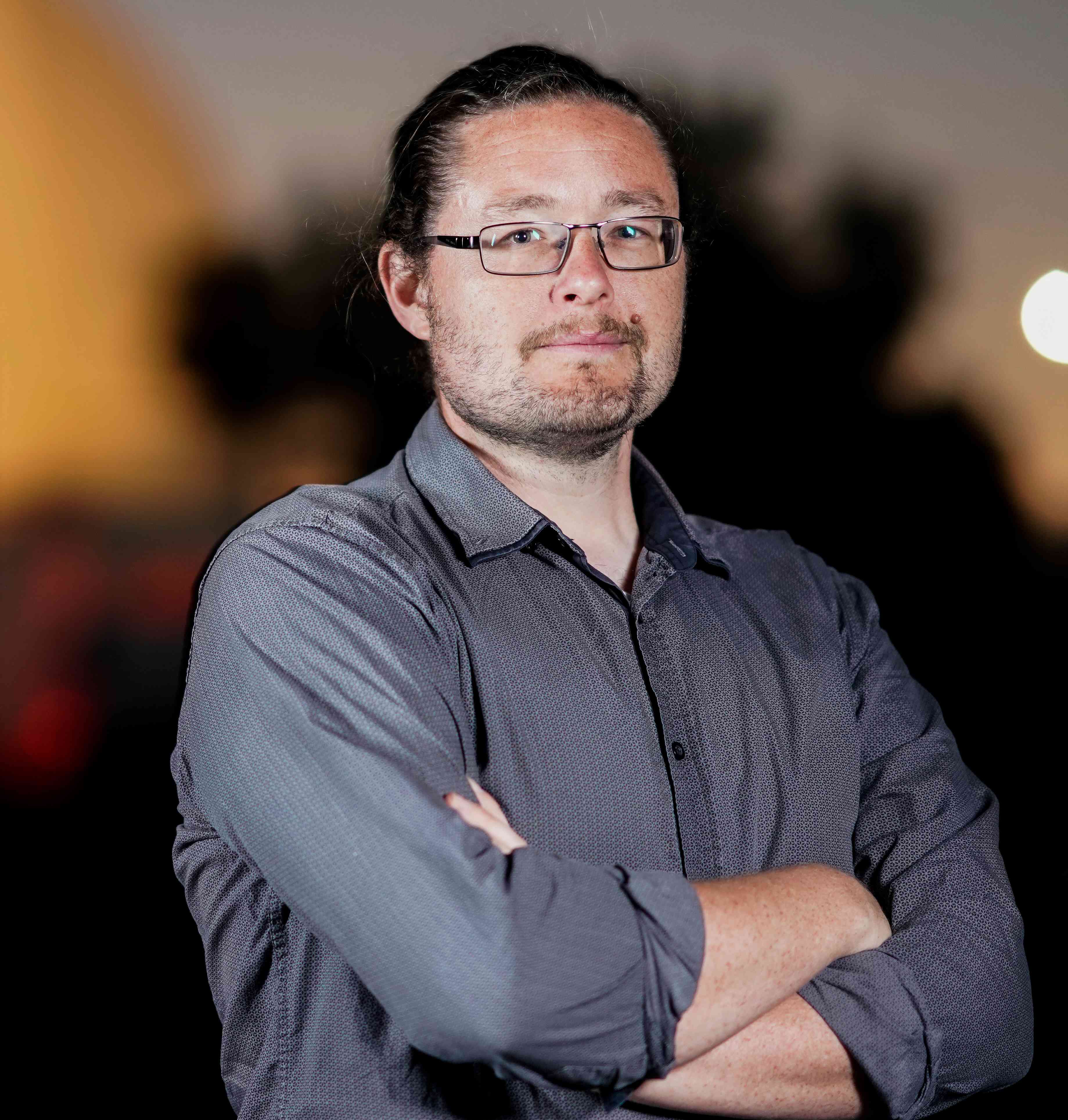 A portrait-flat photo of astrophysicist Brad Tucker, he is standing slightly angled towards the right with with his arm. He has black hair, glasses, a buttoned grey-shirt and a beard.