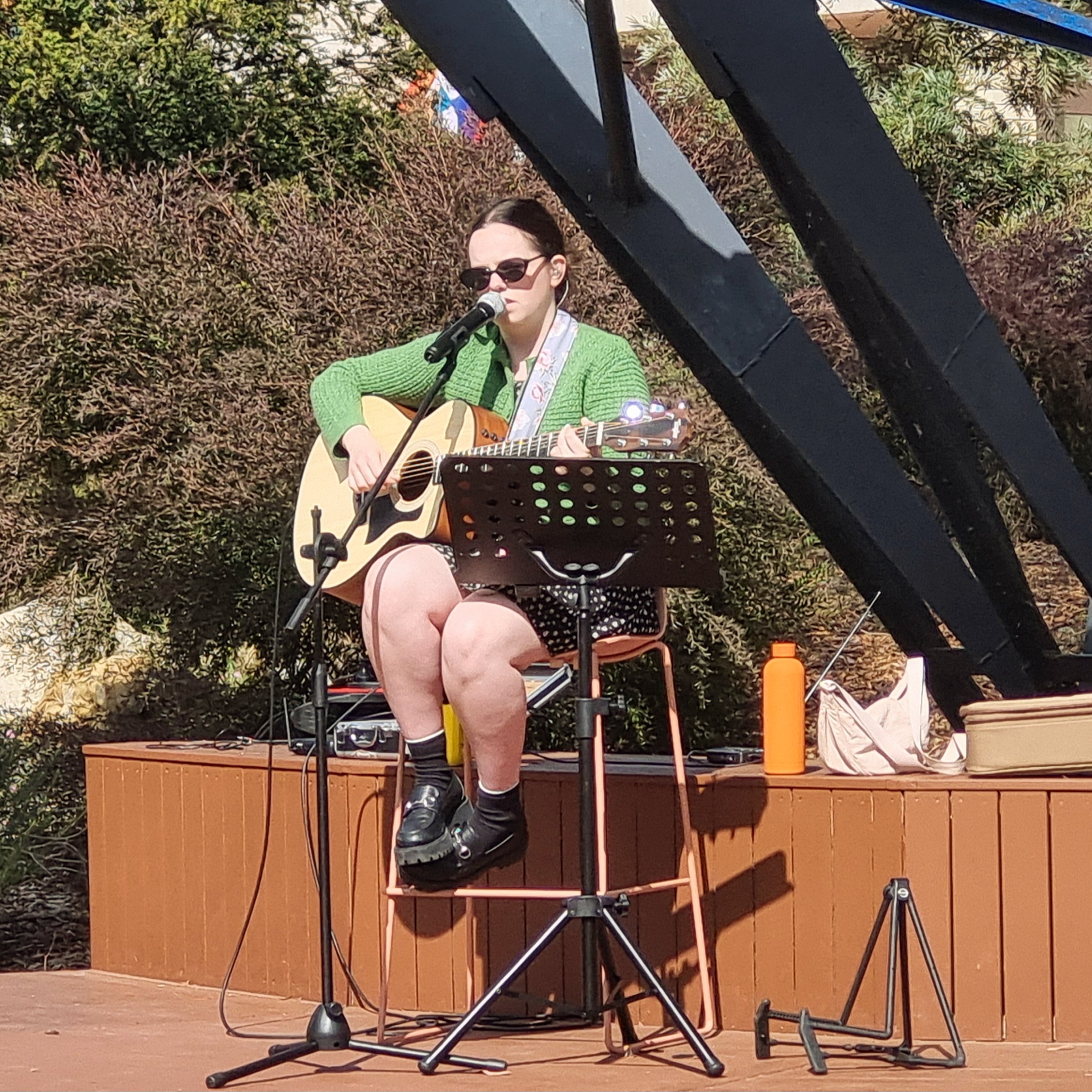 Sophie Edwards, seated on an outdoor stage in the sun, playing guitar and singing.