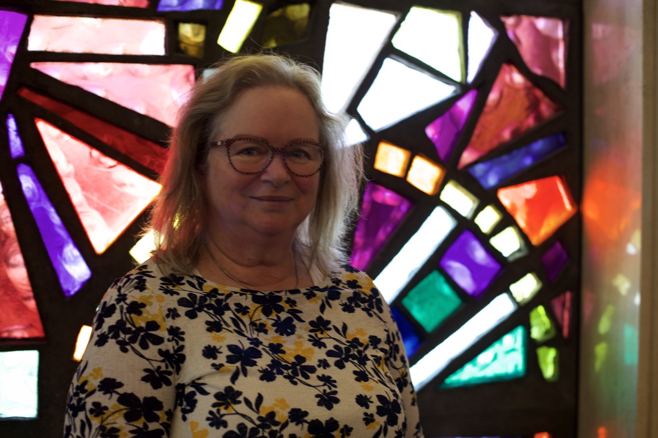 Photo of writer Donna Hanson, infront of stained glass window. She is wearing glasses and has a floral top on. 