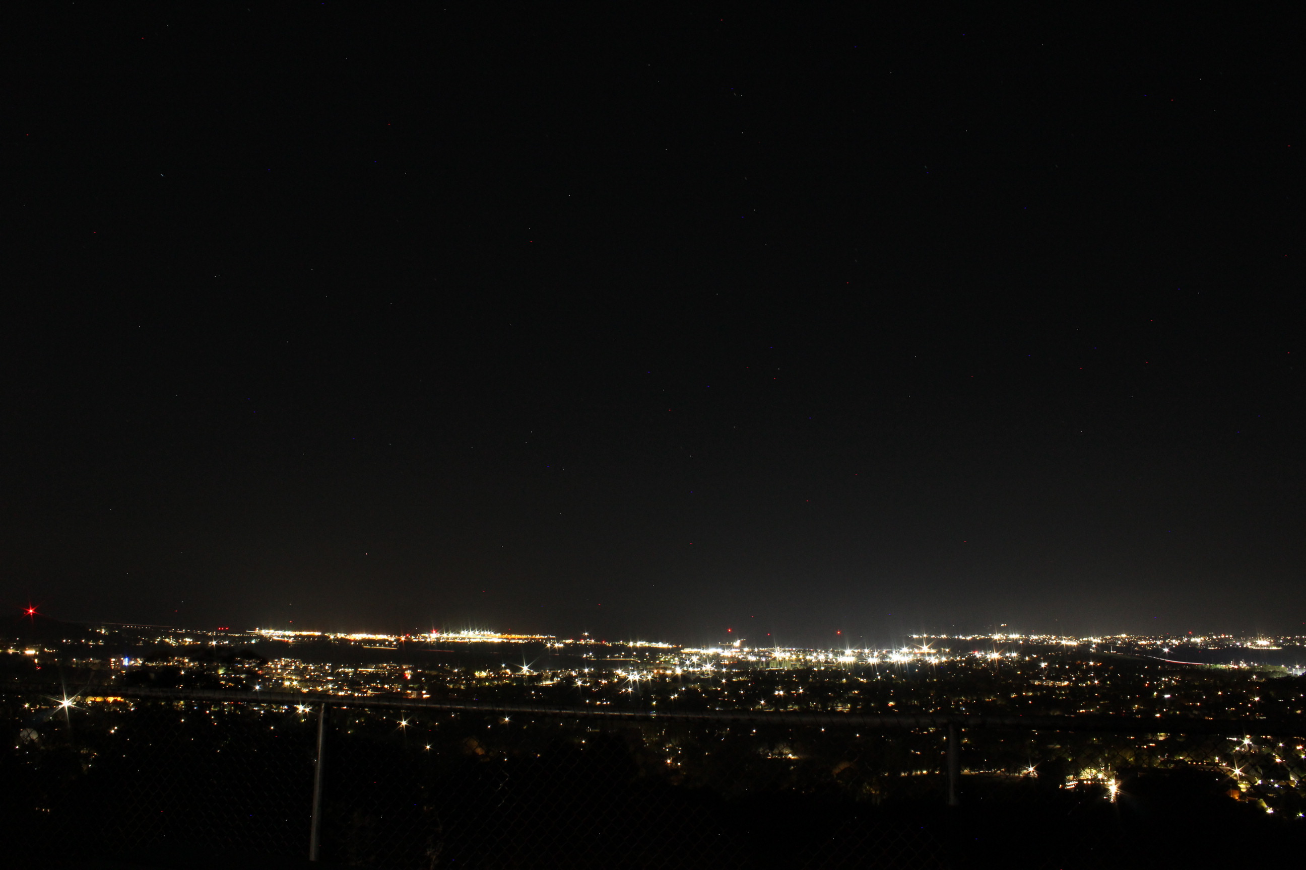 A landscape photo of Canberra at night from Red Hill Lookout. Bright yellow lights pepper the landscape, creating visible sky glow in the background. 