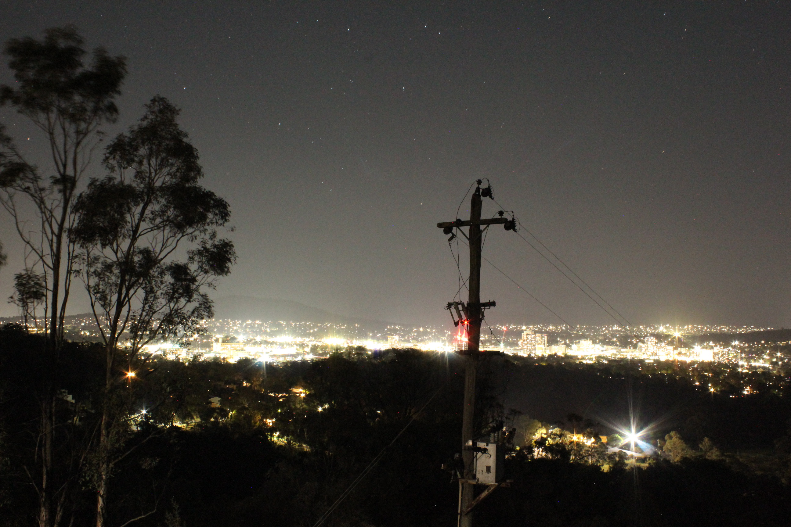 A landscape photo of Canberra at night from Redhill Lookout. A tree and telephone pole sit in the foreground, while the city, trees, and mountains sit in the background. Glaring light from the city flashes towards the viewer and creates prominent sky glow. 