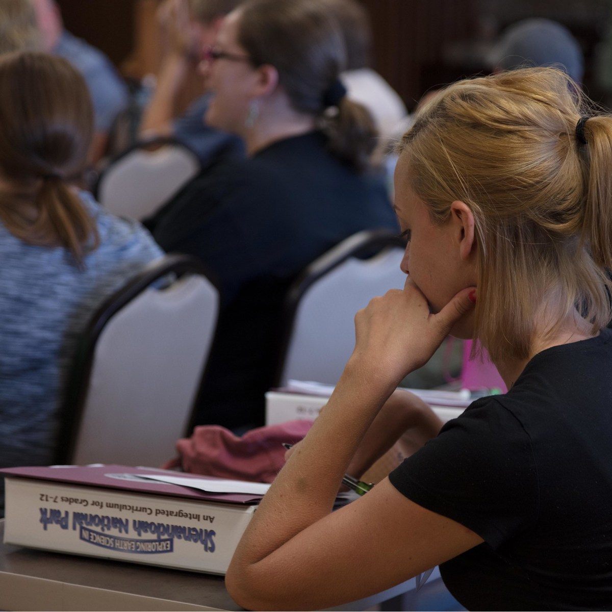 A young teacher sits in a classroom, looking comtemplative. There are other teachers in the backgound. She has a manual on the desk in front of her
