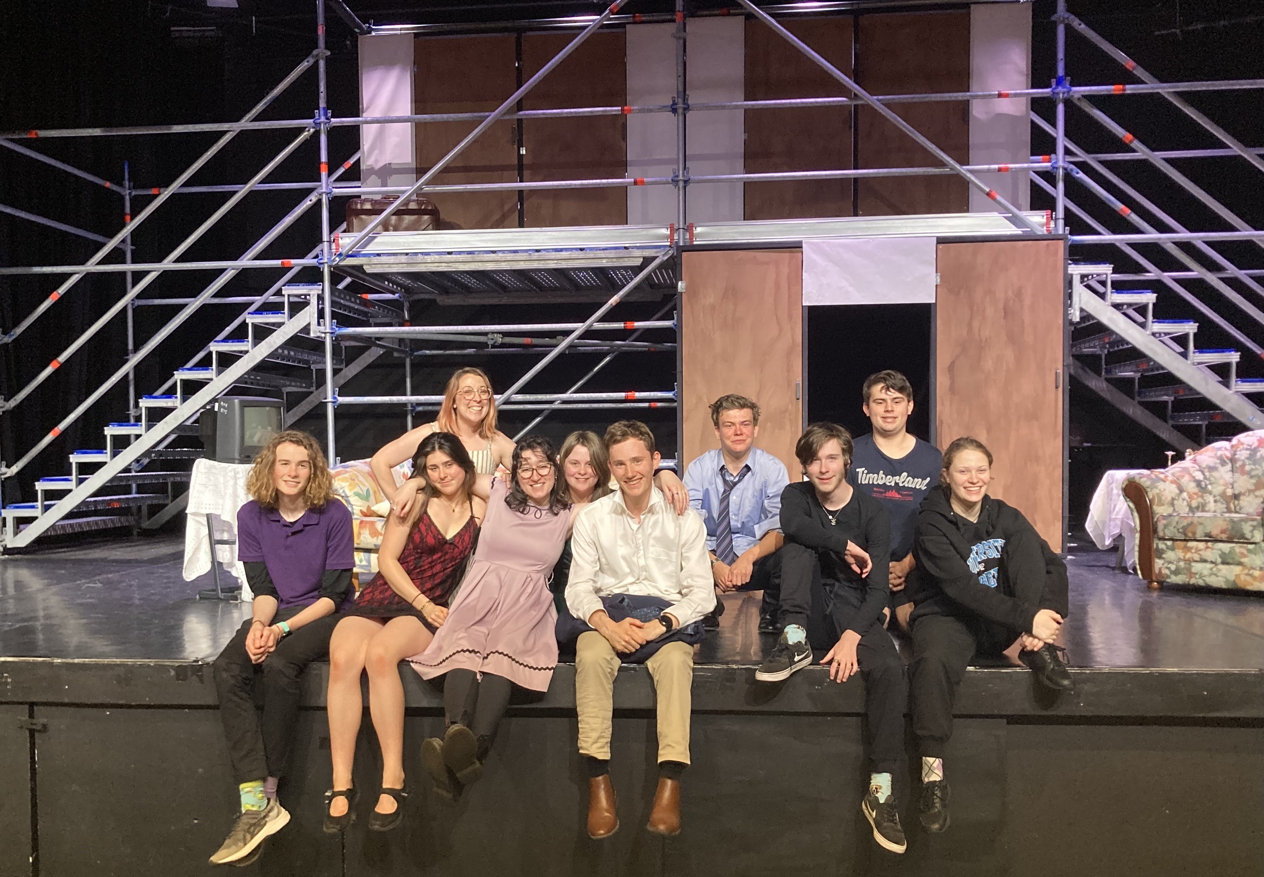 10 people sit on the stage with their arms around each other smiling.