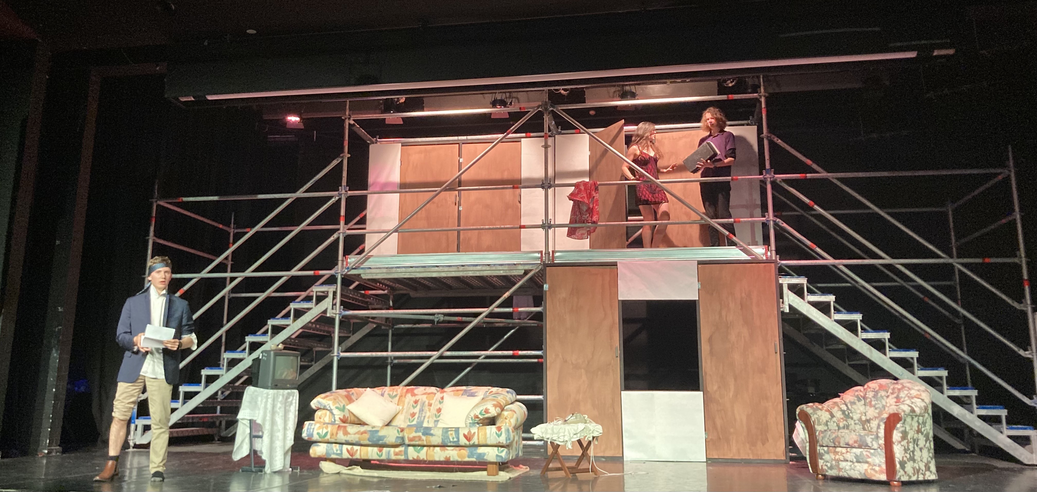Three actors on stage - one is standing on the floor and two are at the top of a scaffolding balcony