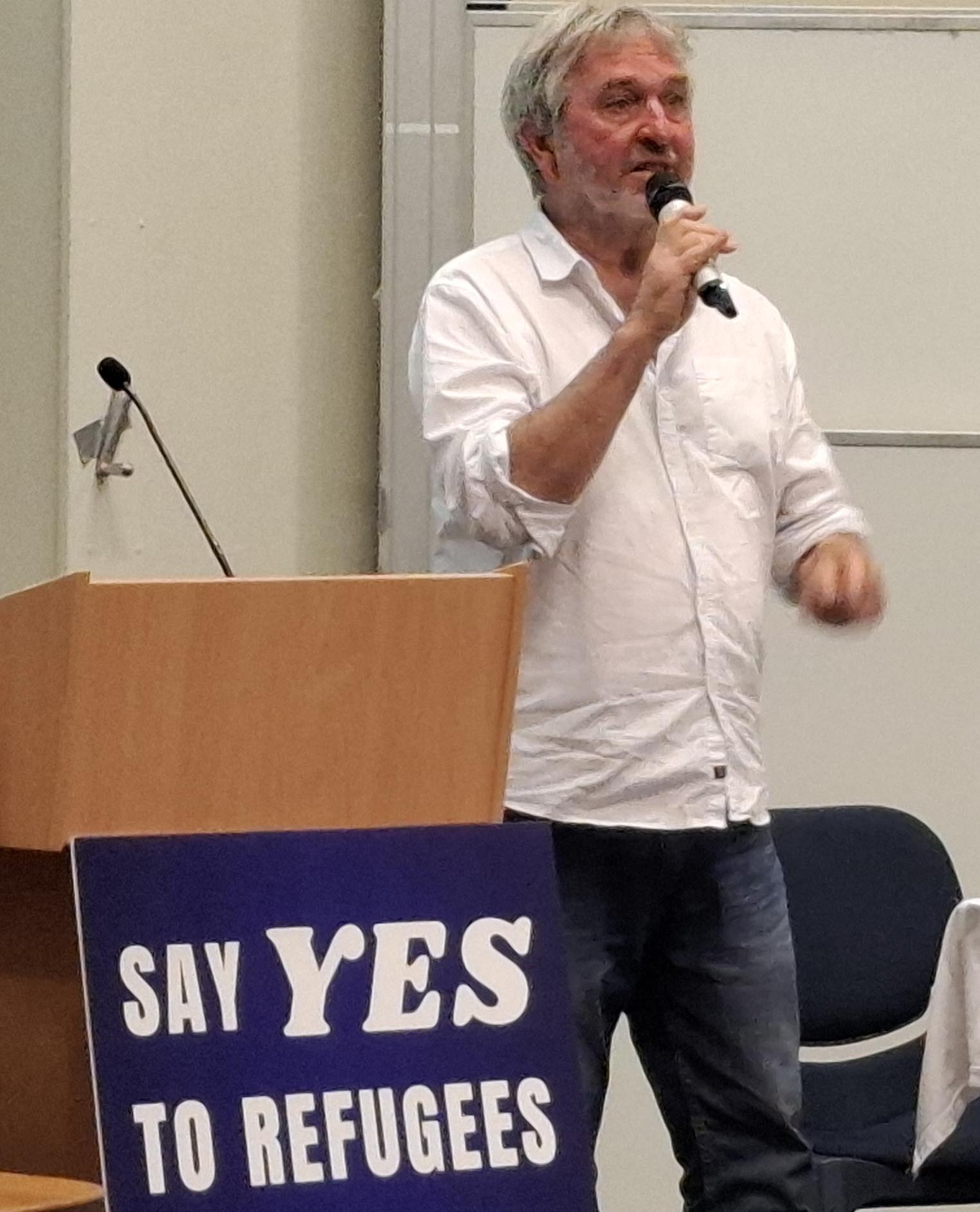 Professor John Minns answering questions in front of a Say YES to Refugees poster