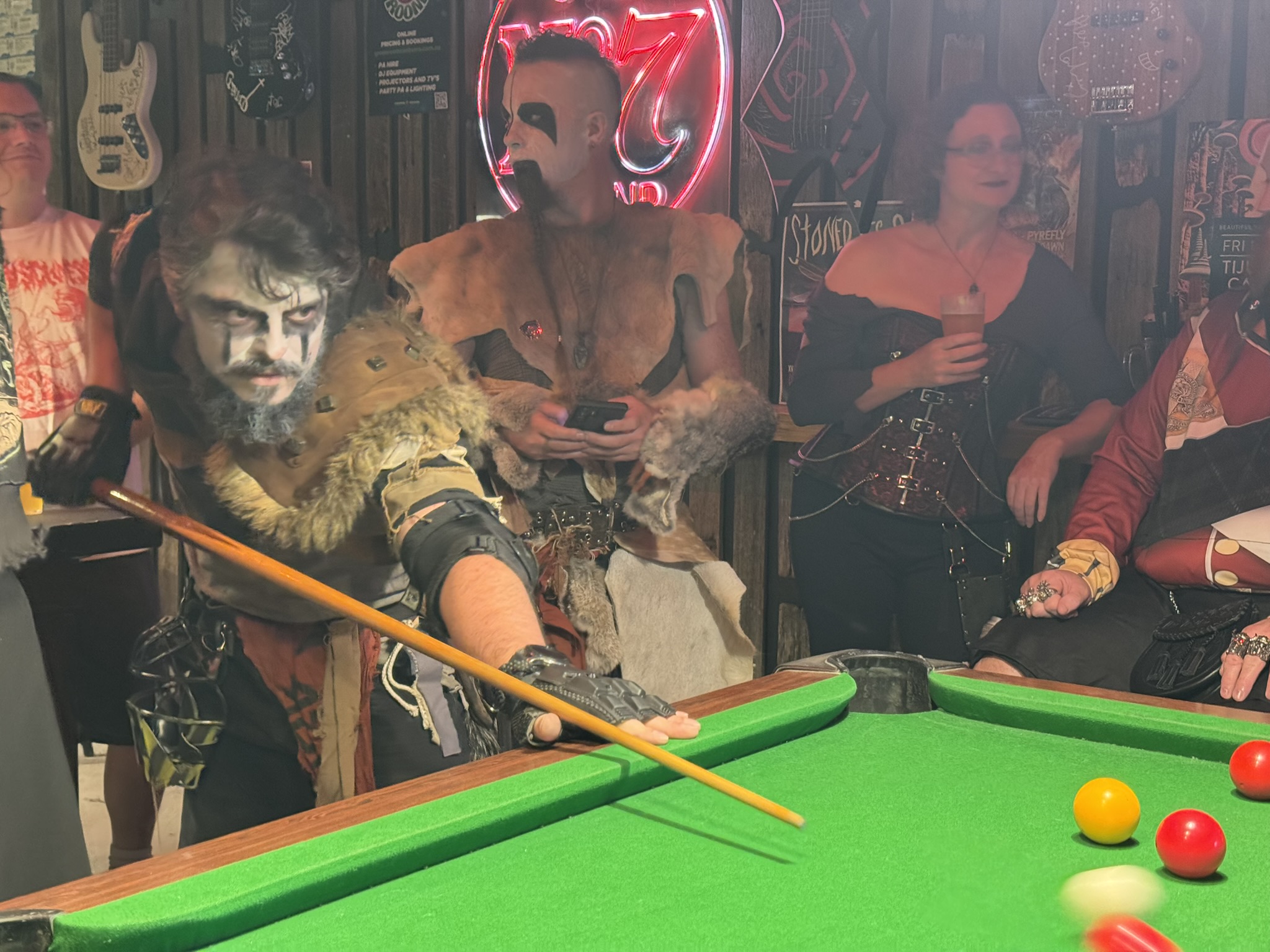 A man in white facepaint and a fur outfit plays pool
