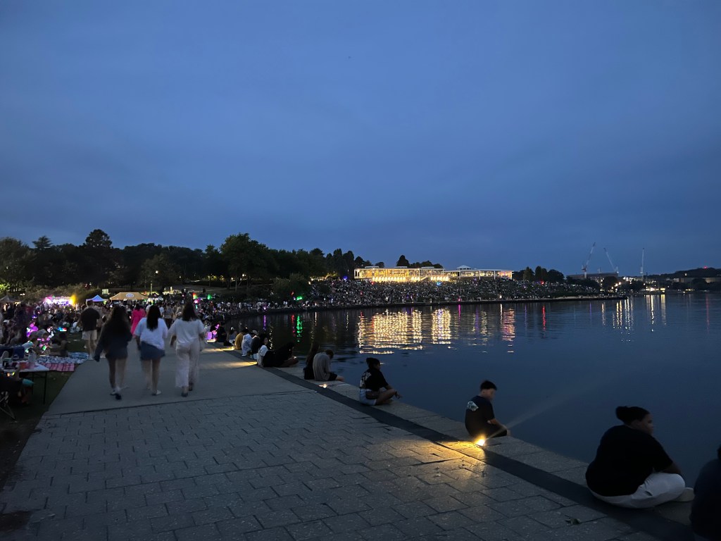 A crowd of people gathering by lake burley griffin with the national capital exhibition building in the background