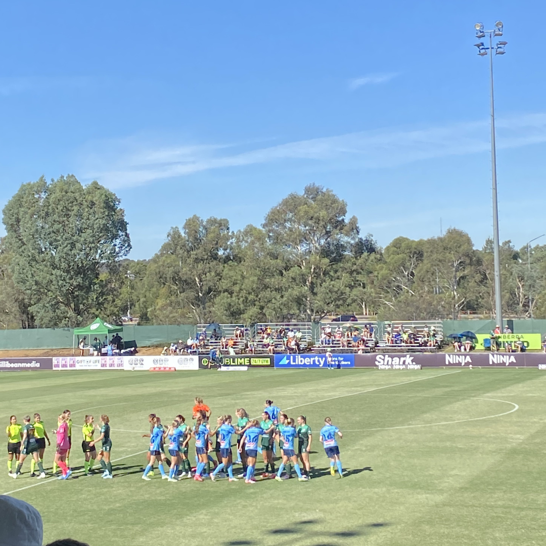 Sydney FC and Canberra United shake hands in a sun-soaked McKellar park, with plenty of fans in the stands behind them, just minutes before kicking off the match.